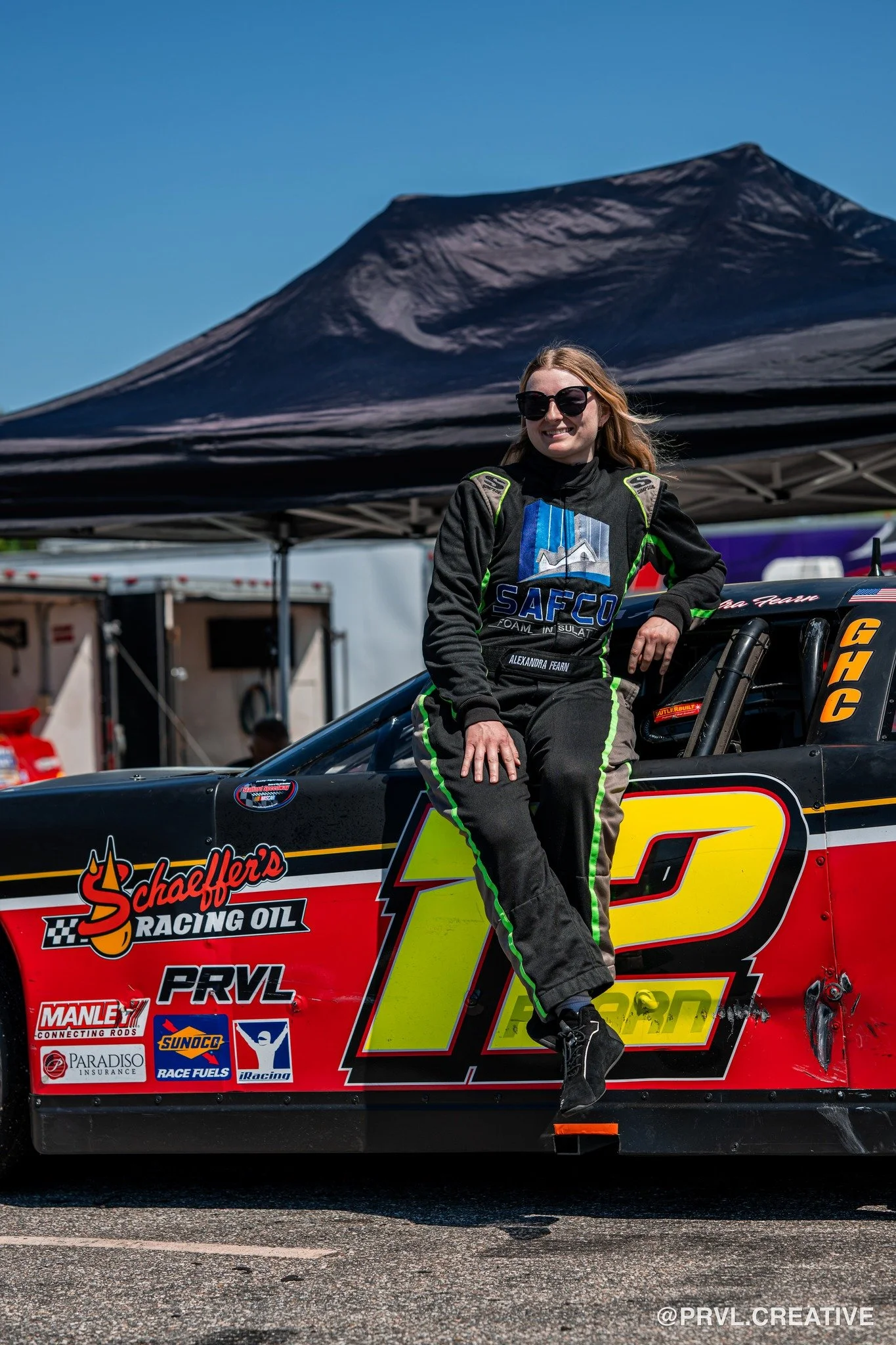 A woman in a racing suit sitting on the side of a race car under a black canopy on a sunny day.