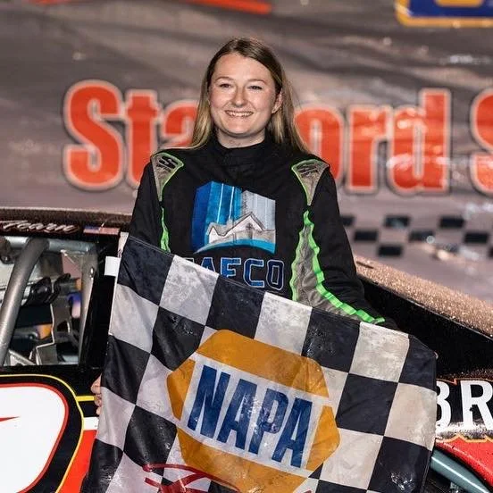 A woman in a racing suit holding a checkered flag with a NAPA logo, standing next to a race car in a racing track, smiling after a race.