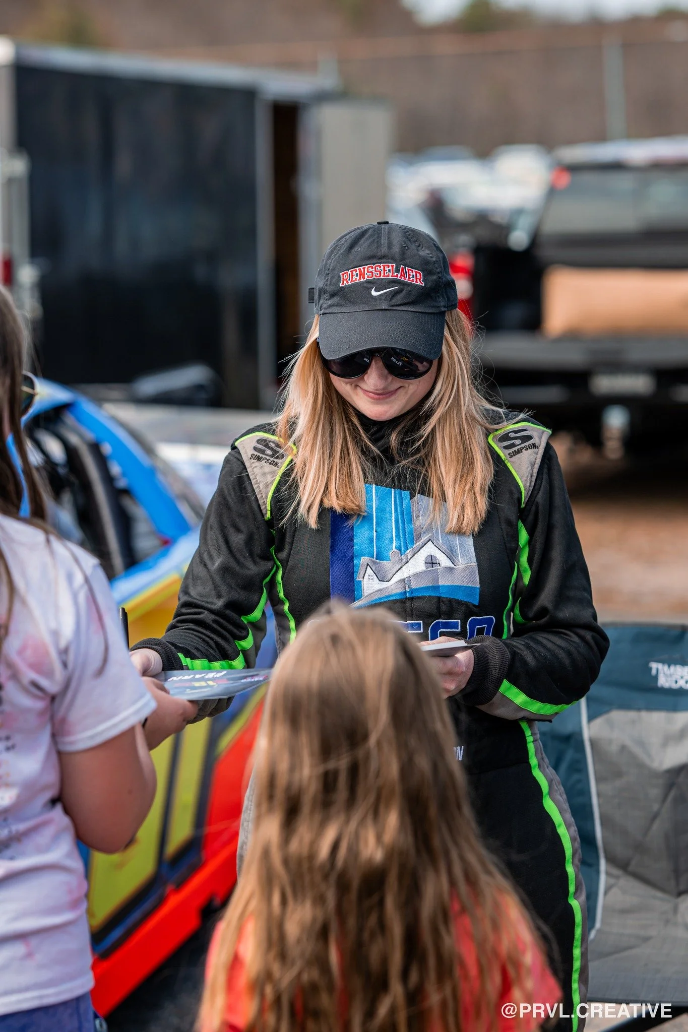 A woman wearing a black racing suit with green accents, a black cap with red text, and sunglasses is signing autographs for children at a race track. Racing cars and trailers are visible in the background.