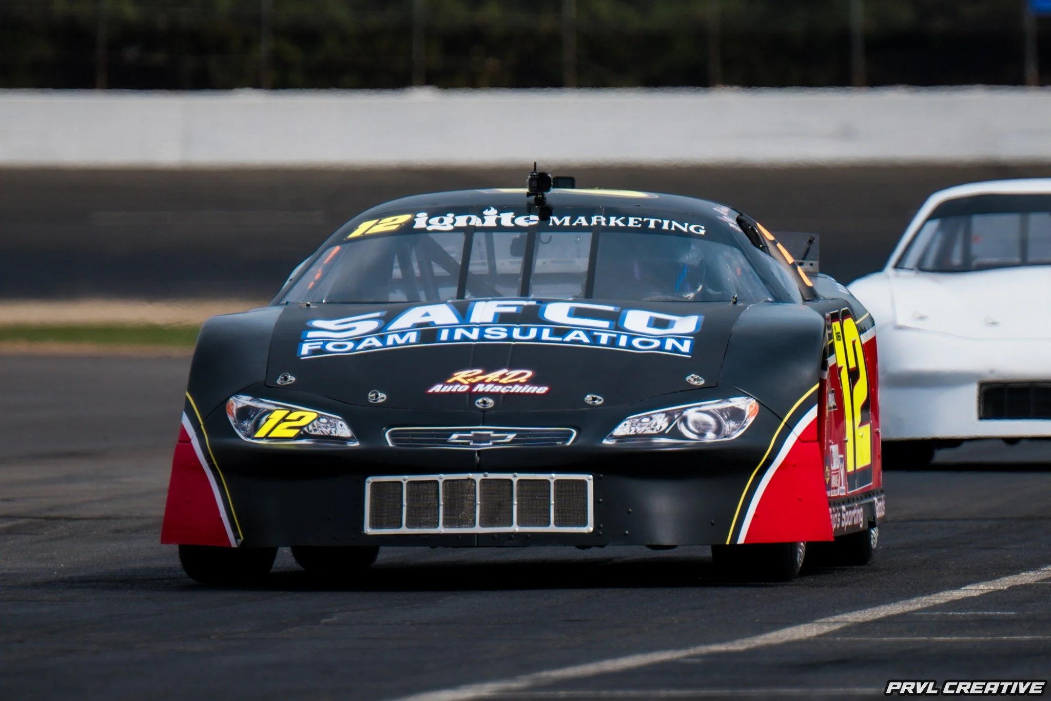 A black race car with yellow and red accents, number 12, on a racetrack, with another white race car in the background.