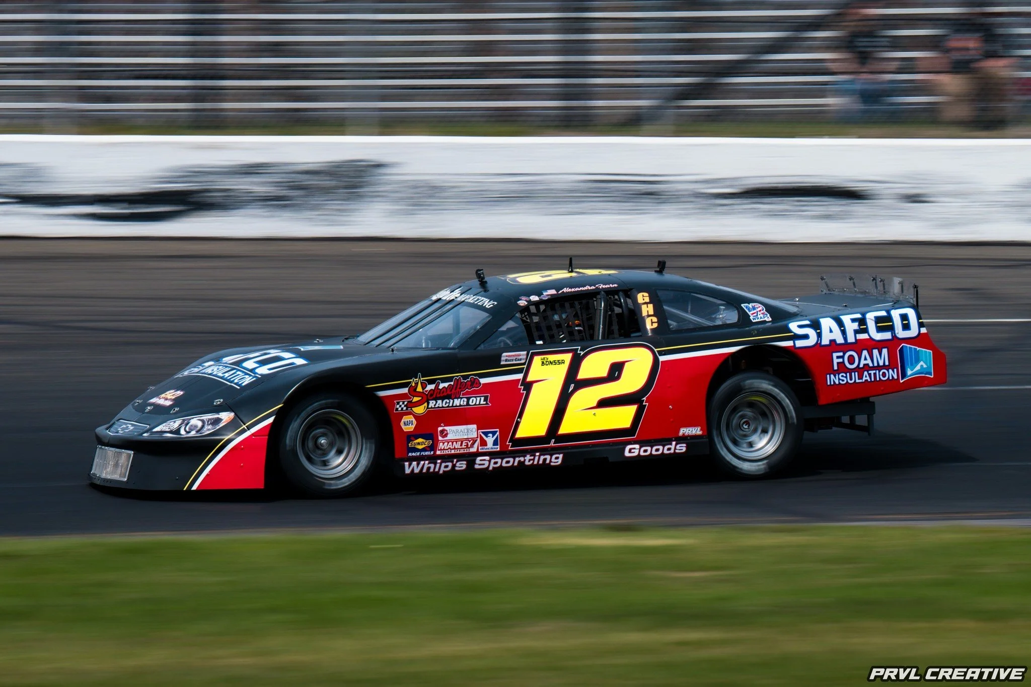 Race car on a track, black with red and yellow accents, number 12, sponsored by Safco Foam Insulation with various logos, driven by Alexander George.