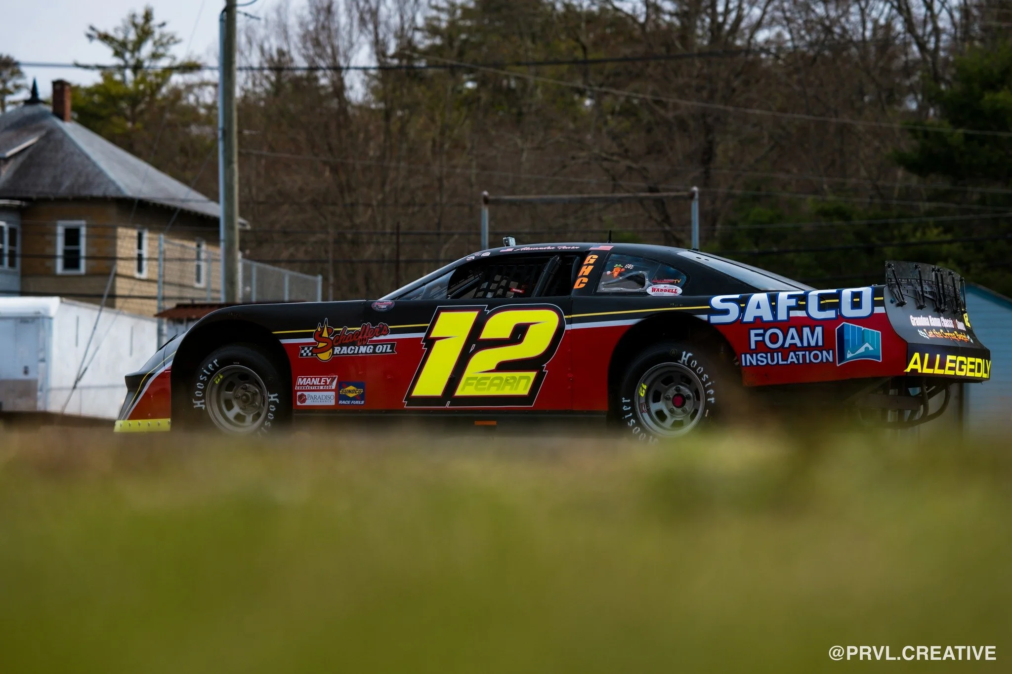 Race car with black, red, and yellow paint, number 12, on a grassy field with a house and trees in the background.