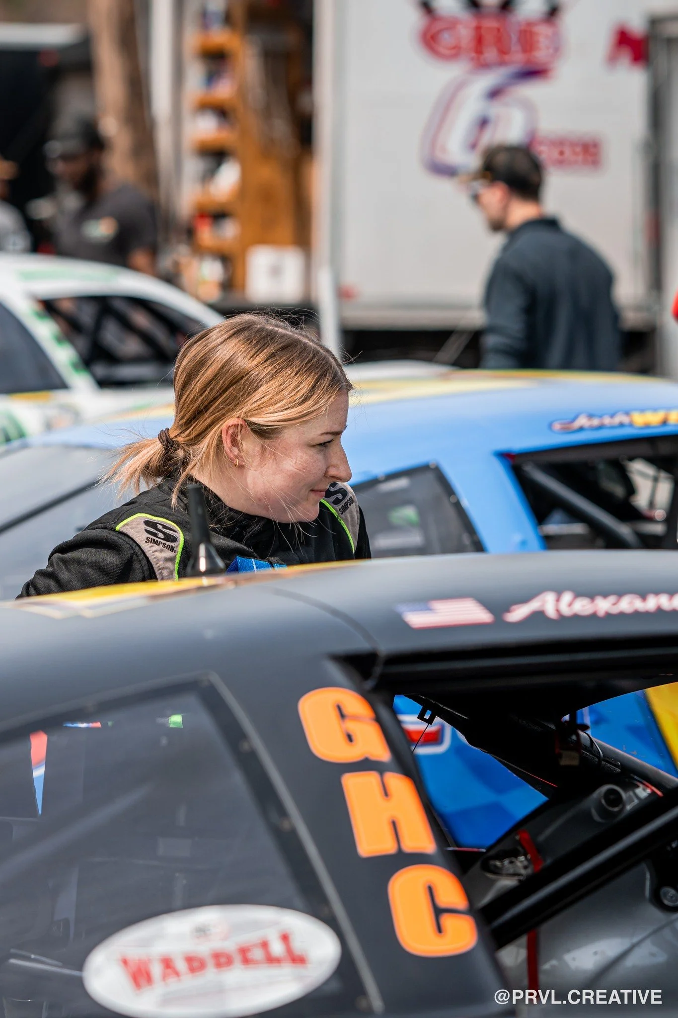 A female race car driver with blonde hair tied back, wearing a racing suit, is smiling as she leans over her race car in a pit area. Several race cars and a person in the background are visible.