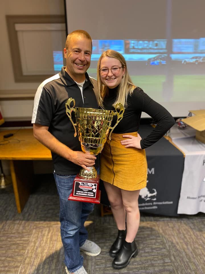 A man and woman smiling and holding a large gold trophy at an indoor event with a screen behind them showing a race track view.