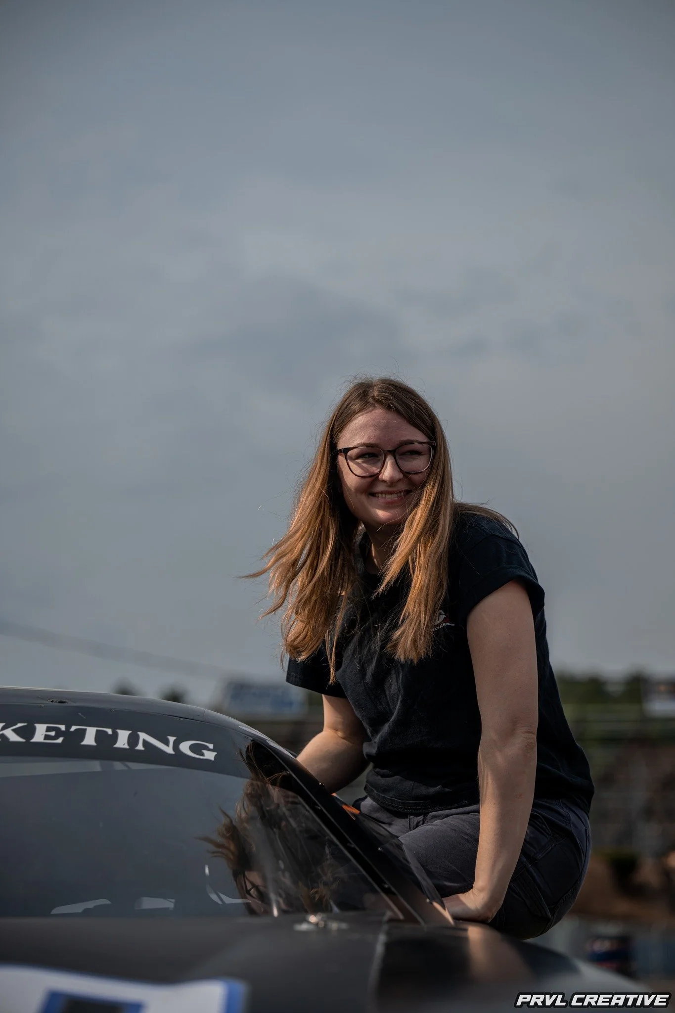 Young woman with long red hair and glasses smiling while sitting on a black car with the word 'MARKETING' visible on the windshield, outdoors on a cloudy day.