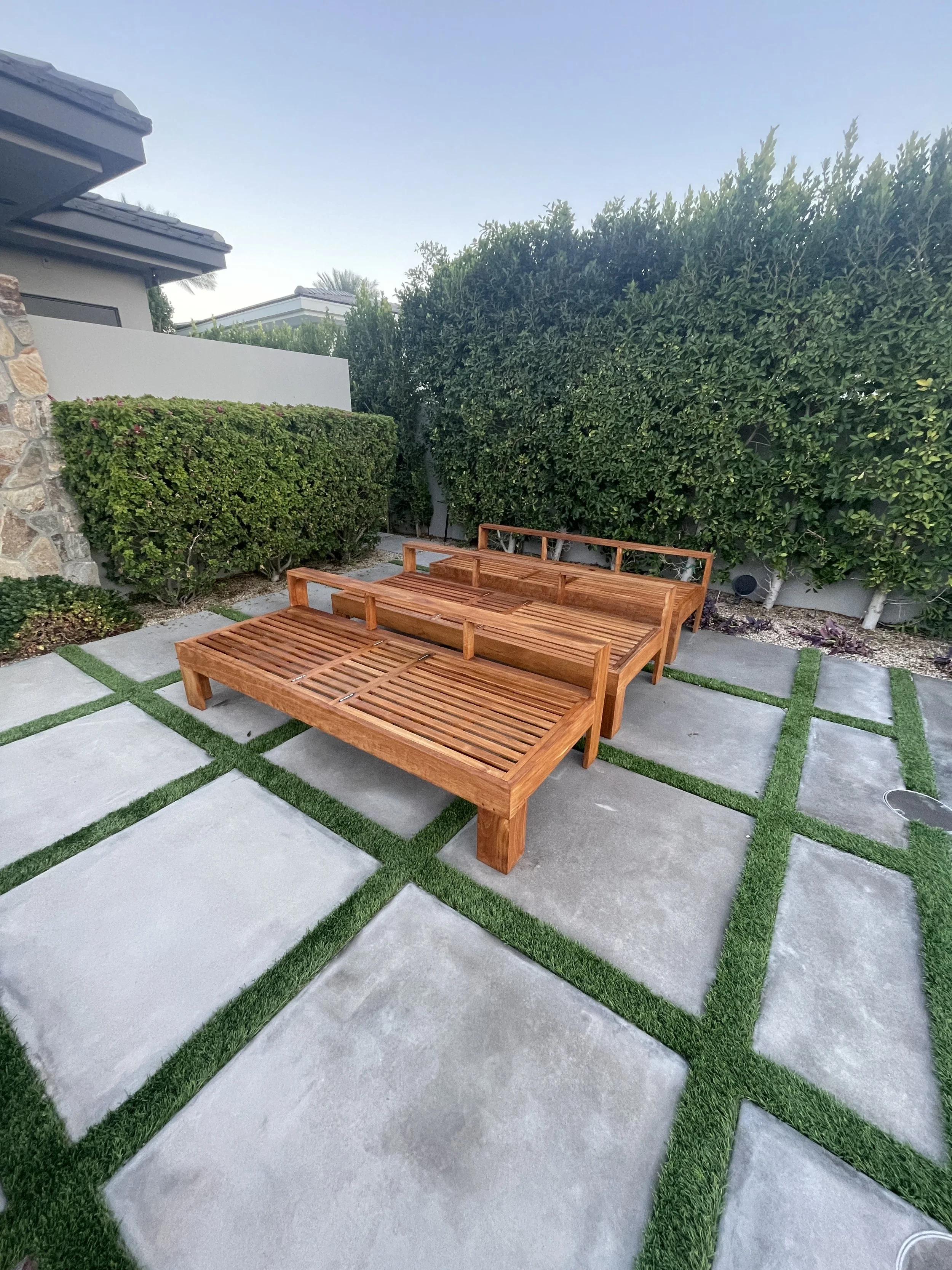 A backyard patio with concrete pavers and green turf accents, featuring two wooden lounge chairs and tall green hedges along the perimeter.