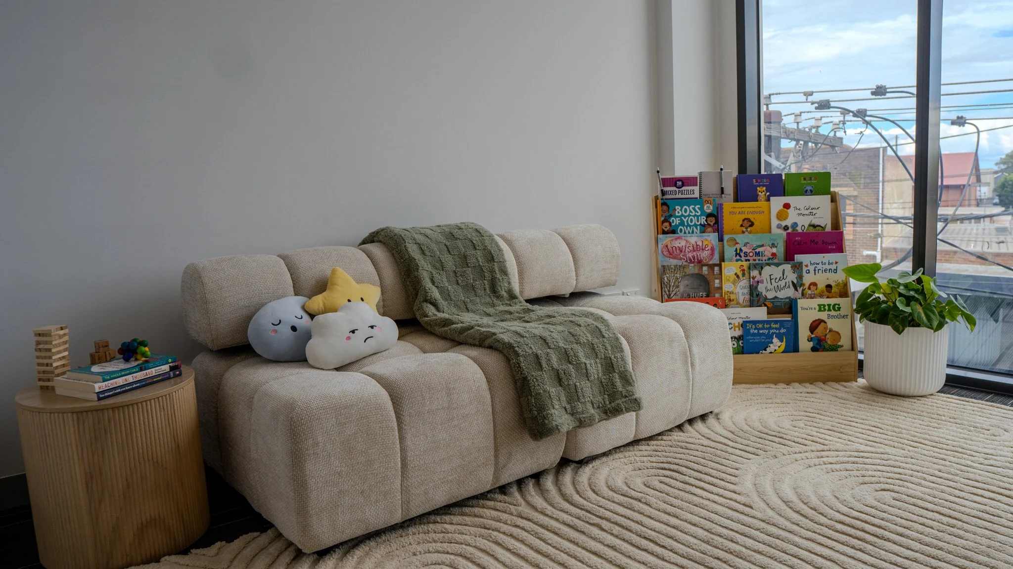 A cozy indoor space with a cream-colored sofa, plush cloud and star pillows, a green blanket, a round wooden side table with children's books and toys, a wooden book display filled with colorful children's books, a large potted houseplant, and a floor-to-ceiling window showing a cityscape with power lines and buildings.