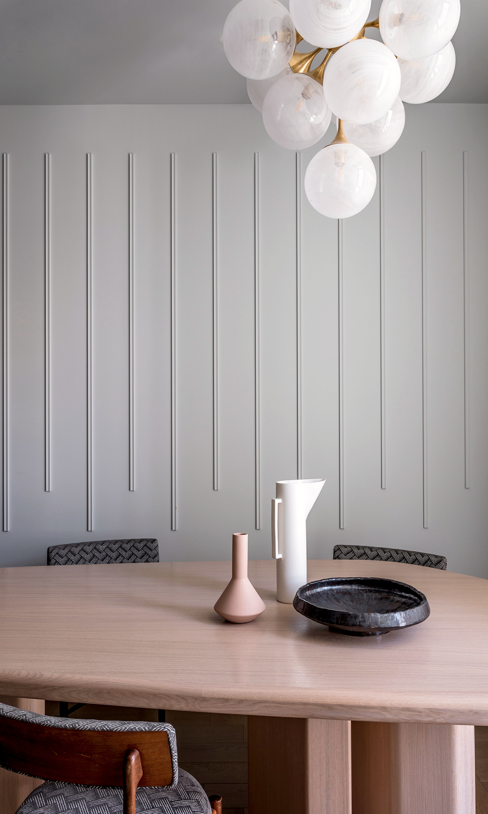 A minimalist dining room with a wooden table, a decorative vase, a pitcher, a ceramic bowl, and a modern chandelier. The room features a textured gray wall with vertical grooves and modern chairs.