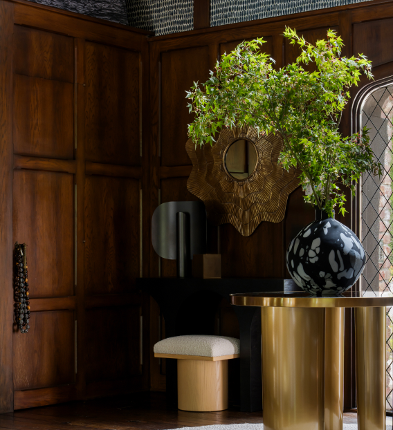 Elegant interior with wood paneling, decorative mirror, marble vase with green plant, modern console table, and upholstered stool.