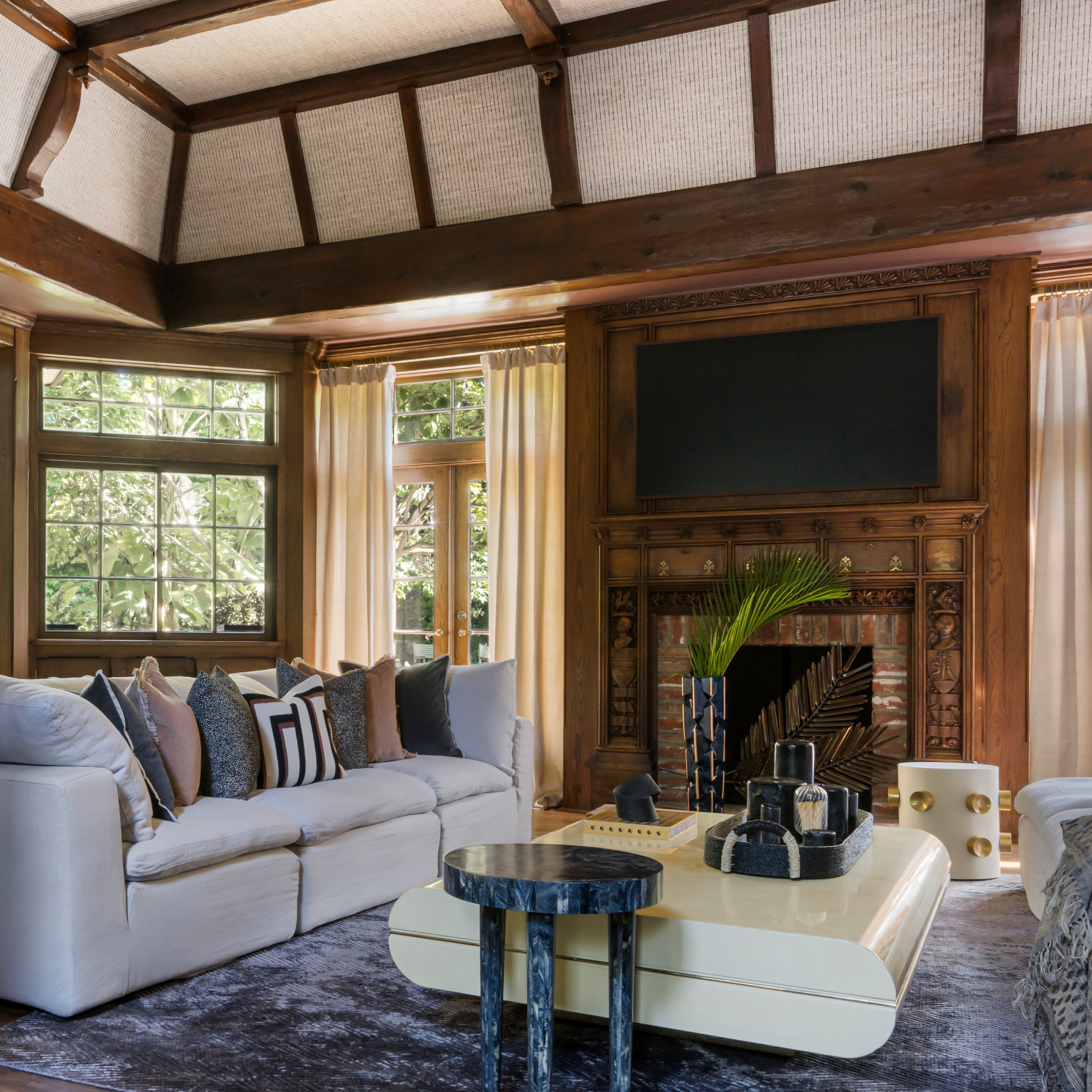 Luxurious living room with a white sofa, patterned cushions, and a modern coffee table. Features a wooden beamed ceiling, large windows with sheer curtains, and a brick fireplace with decorative wood mantling. The room is elegantly decorated with plants and stylish decor accents.