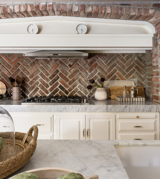 Modern kitchen with brick backsplash, marble countertops, white cabinets, gas stove, and range hood. Decor includes cutting boards, utensils in a holder, a vase with flowers, and a decorative basket.