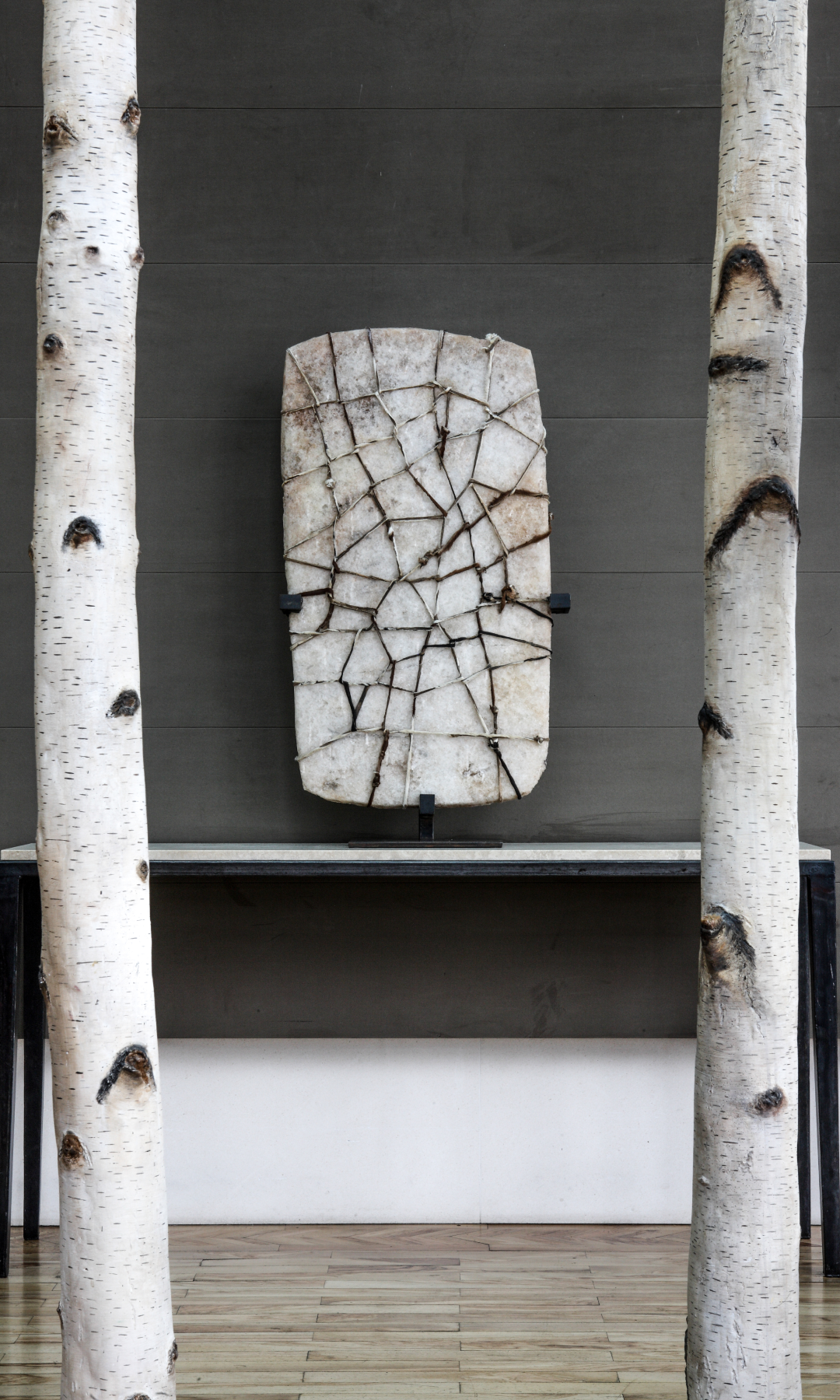 Two birch tree trunks flank a table displaying a cracked stone artwork against a dark wall.