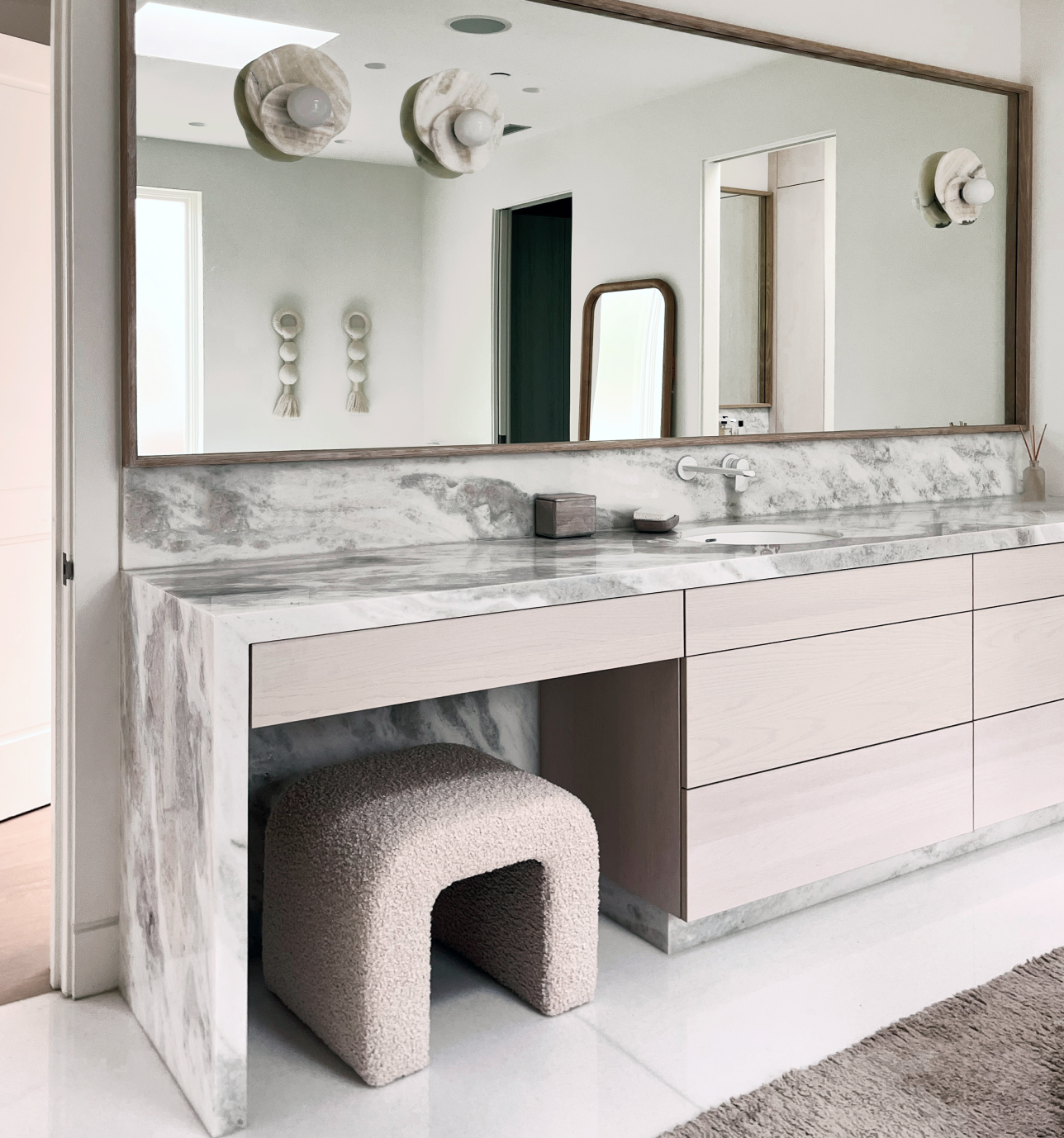 Modern bathroom with marble countertop, large mirror, minimalist decor, and a plush stool underneath the vanity.