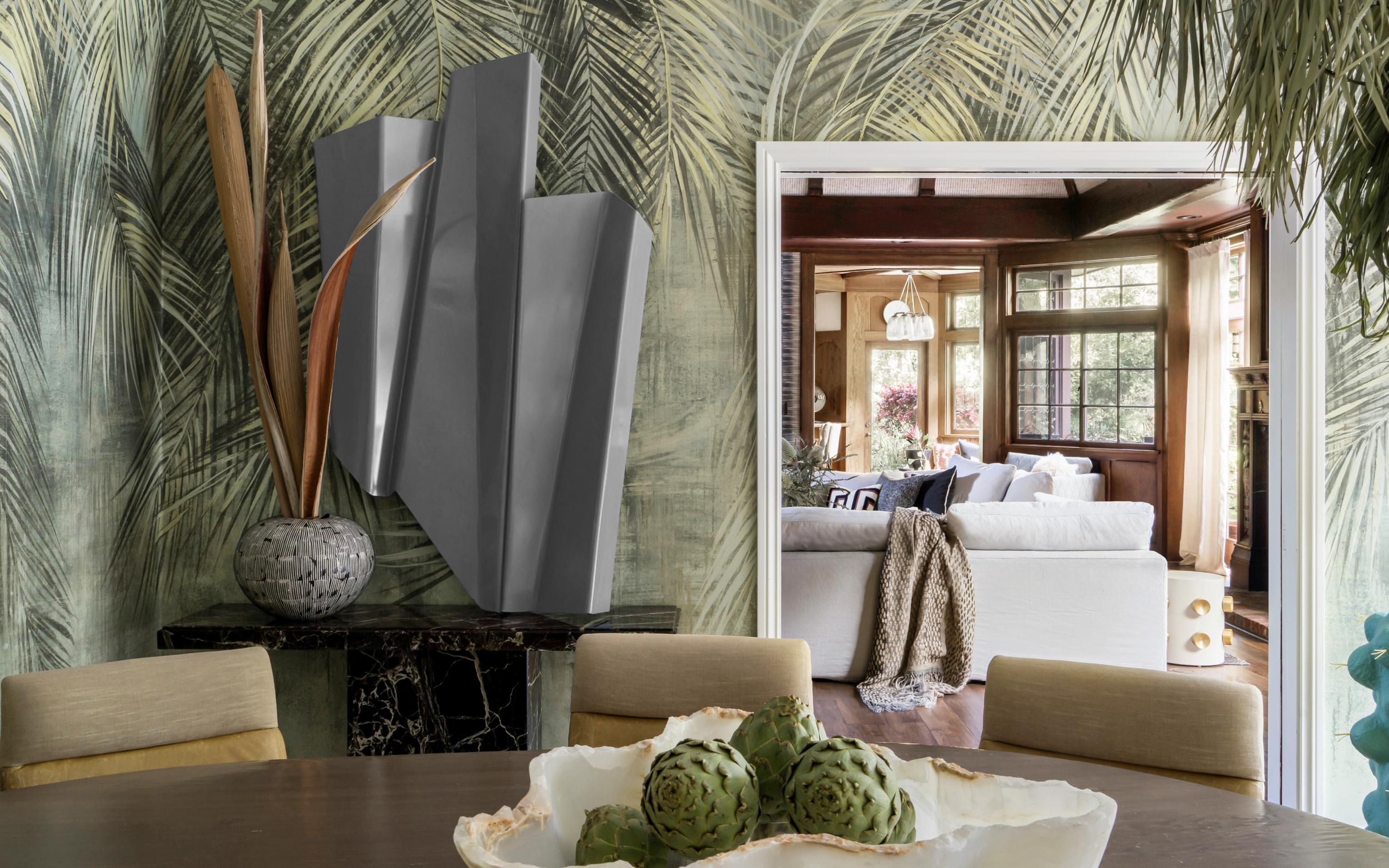 Modern dining area with tropical wallpaper, abstract wall art, decorative vase with leaves, and a view into a cozy living room with large windows and comfortable seating.