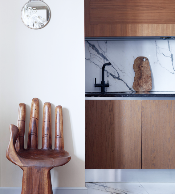 Interior scene with a wooden hand-shaped chair next to a kitchenette featuring a marble backsplash, wooden cabinets, and black faucet. A small circular mirror is on the wall.