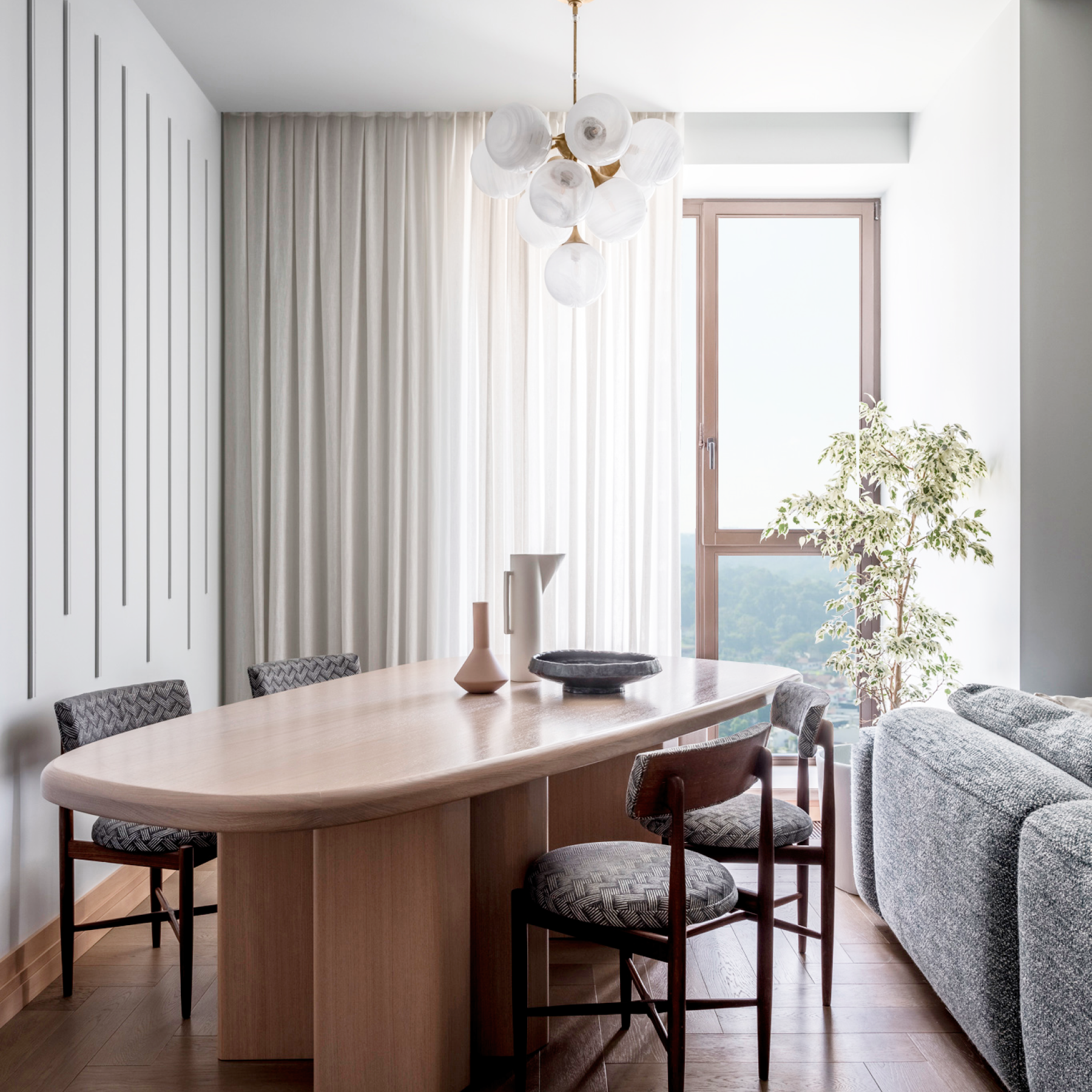 Modern dining area with a wooden table, upholstered chairs, decorative vases, and a round light fixture. Large window with sheer white curtains and a potted plant in the corner.