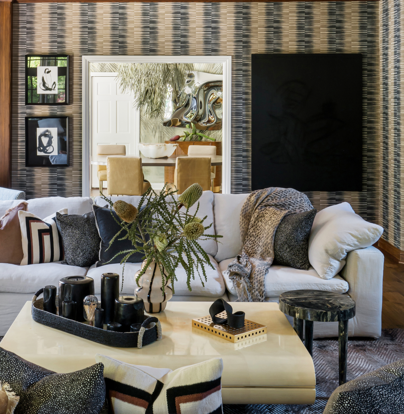 Modern living room with white sofa, patterned pillows, and a decorative vase on a beige coffee table. Geometric wallpaper and contemporary art adorn the walls. Dining area visible in the background.