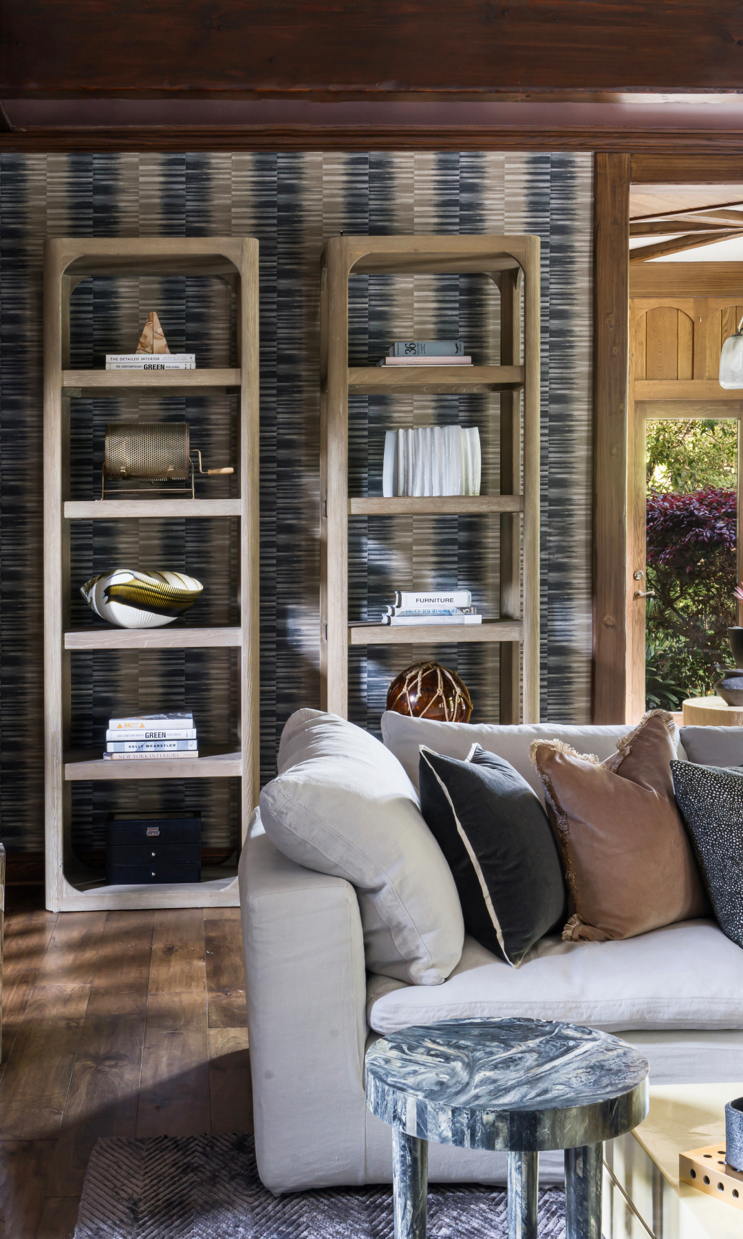 Modern living room interior with two wooden bookshelves against a patterned wall. The shelves are decorated with books, vases, and decorative items. A plush sofa with an assortment of pillows and a small round marble table are in the foreground.