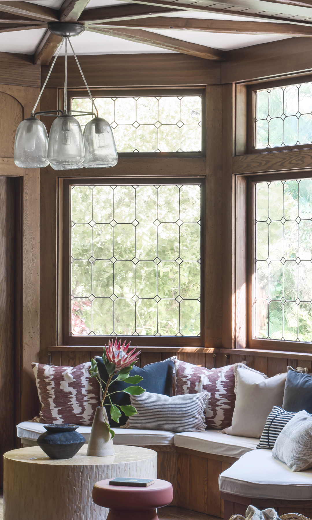 Living room corner with stained glass windows and a cozy seating area. Features a pendant light, cushioned bench with decorative pillows, a round wooden table, and a plant in a vase.