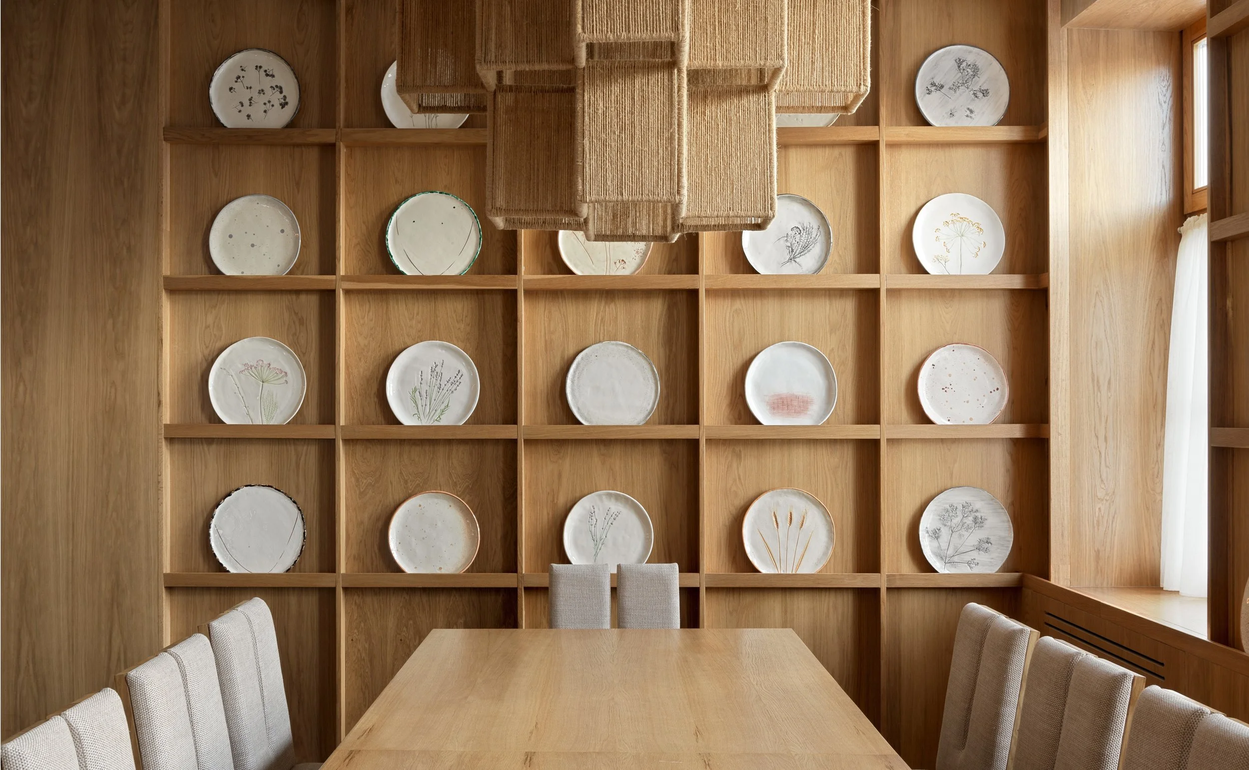 Dining room with wooden shelves displaying decorative plates, a wooden table, upholstered chairs, and a pendant light made of woven material.