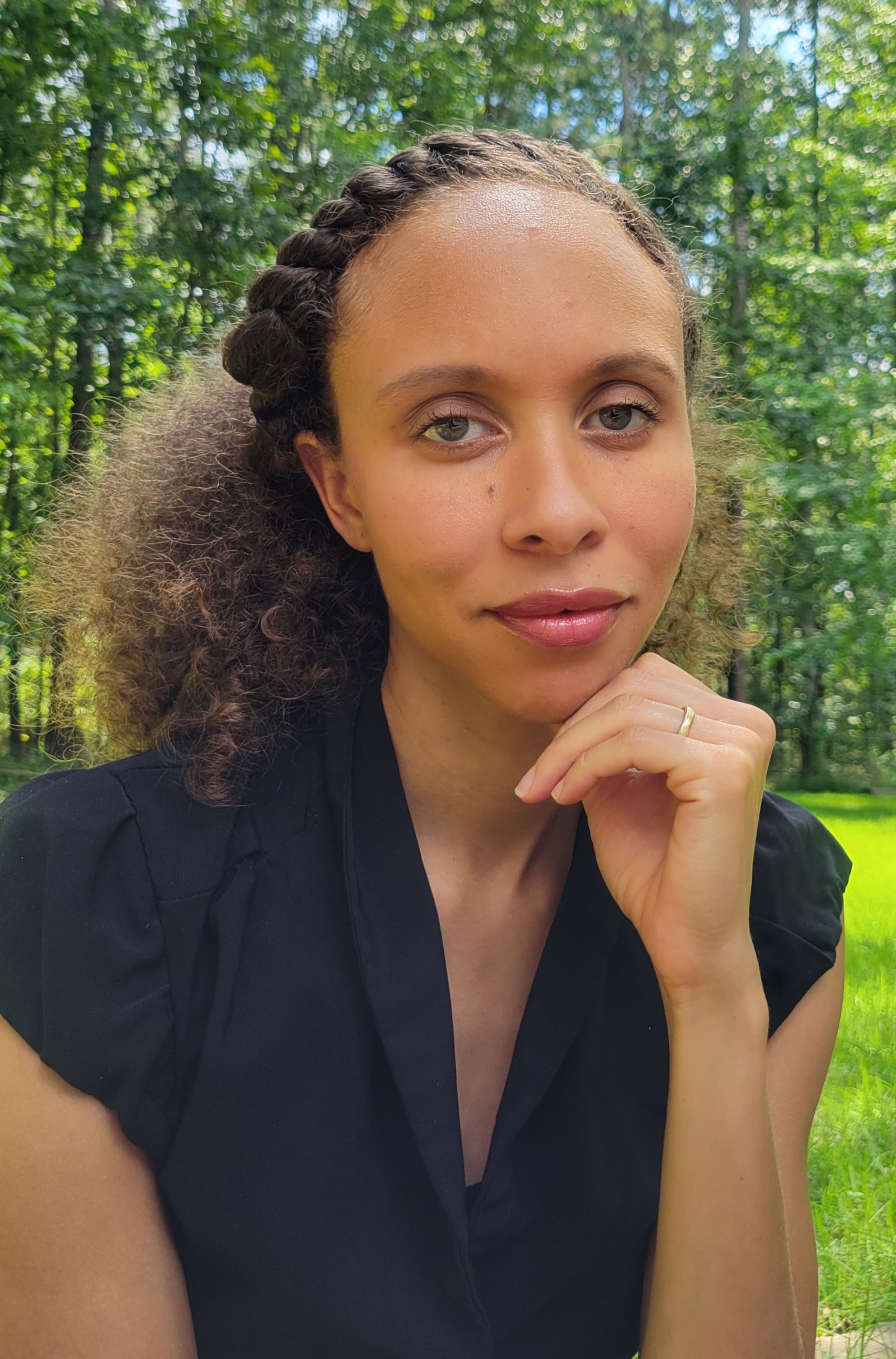 A young woman with curly hair and light skin, posing outdoors in front of green trees, resting her chin on her hand, wearing a black top.