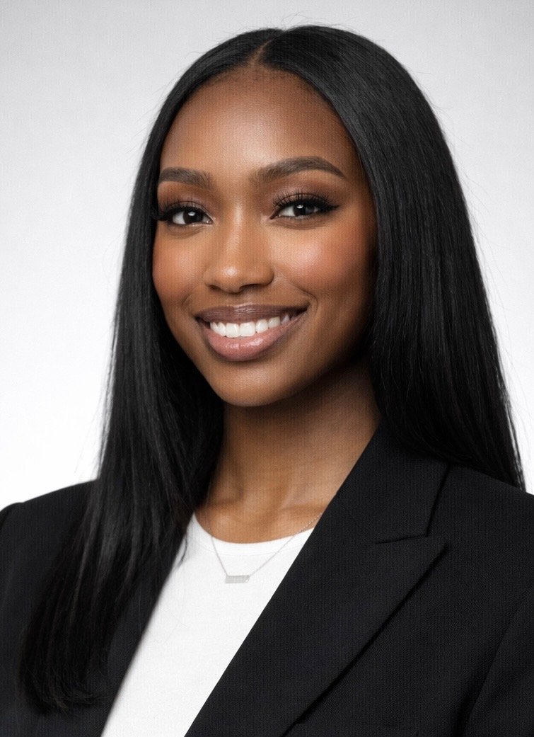 Portrait of a man in a beige suit and blue tie outdoors with green trees in the background.