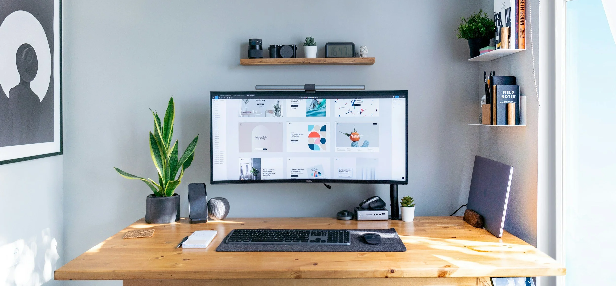 A tidy home office with a wooden desk, large monitor displaying a design website, potted plants, books, and organized shelves on a light gray wall.