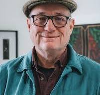 A man wearing glasses, a black hat, a herringbone coat, and a maroon shirt taking a selfie at an airport or similar indoor location.