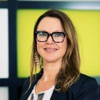 A woman with long brown hair, wearing glasses and a dark blazer, smiling in front of a colorful background.