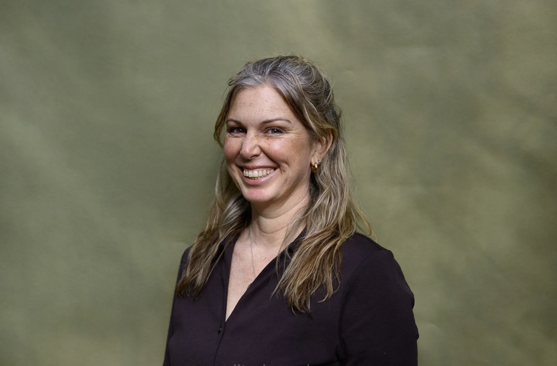 A woman with long wavy hair, wearing a dark blazer, smiling, standing against a plain green background.
