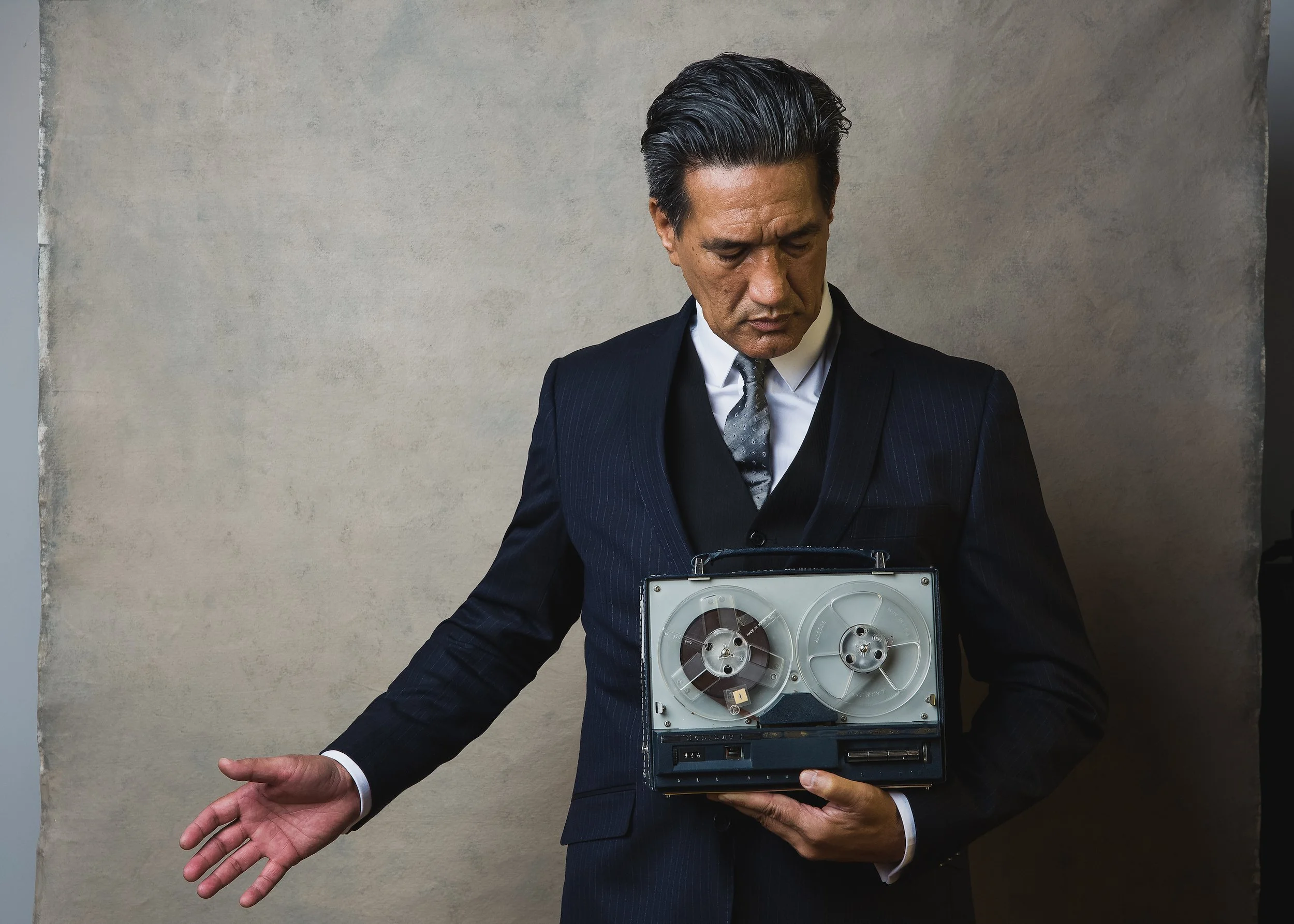A man dressed in a black suit with a white shirt and a tie, holding a vintage tape recorder while standing against a plain wall.