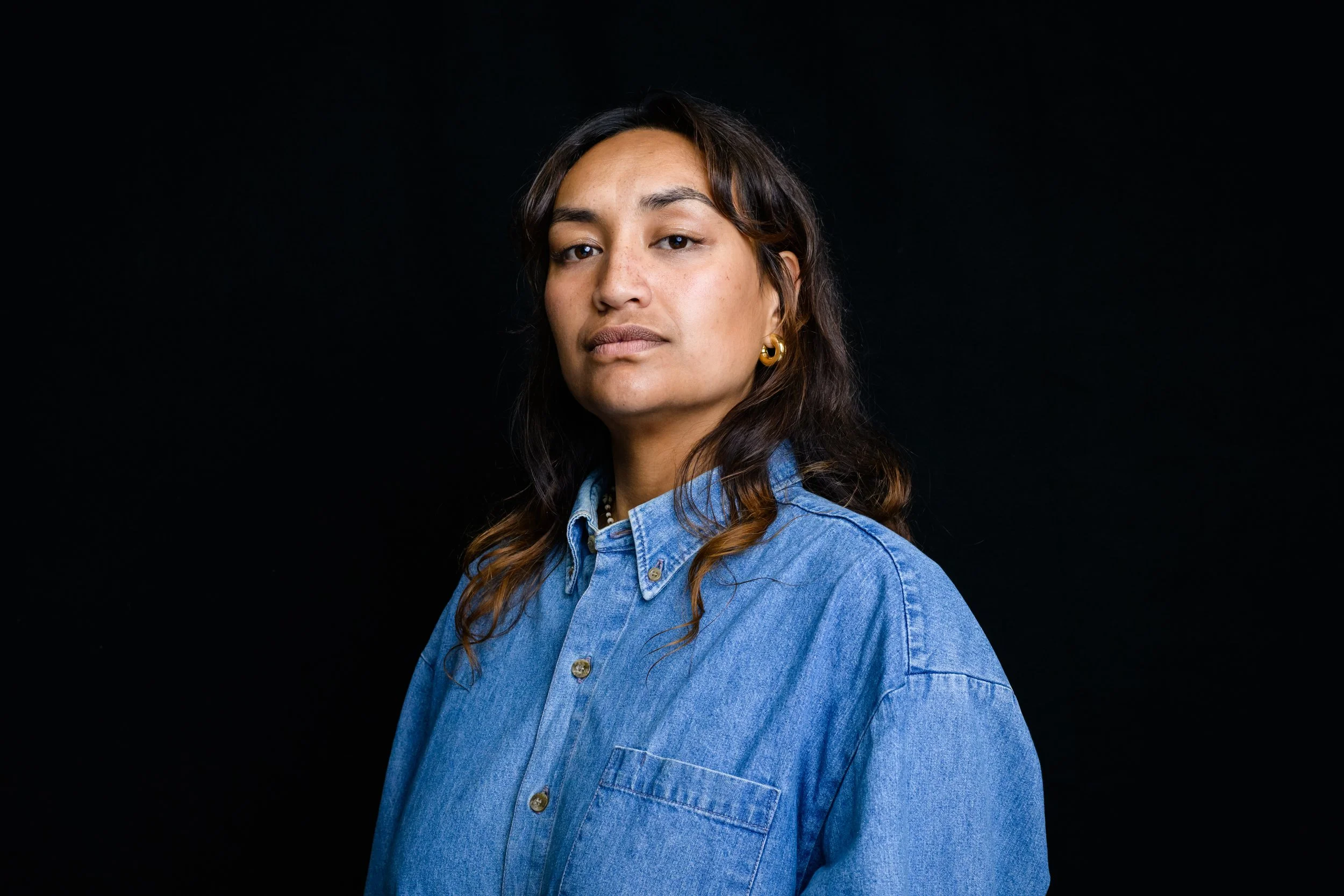 A woman with long dark hair, wearing a denim shirt and gold hoop earrings, looking confidently at the camera against a black background.