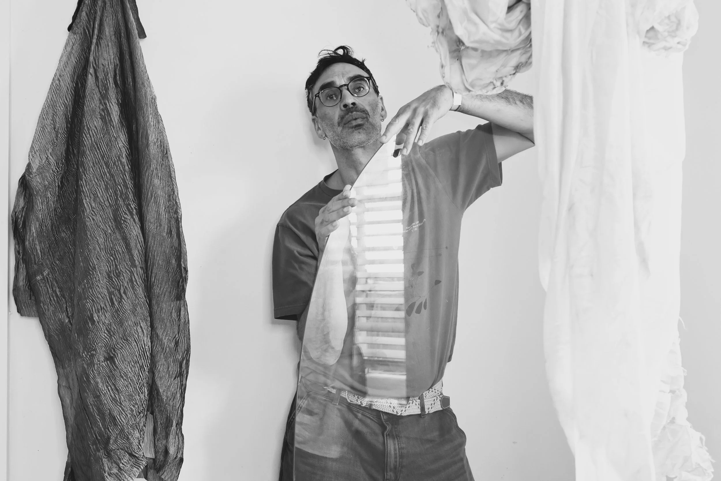 Black and white photo of a man with glasses and earrings holding a mirror, looking at his reflection while standing near hanging clothes. The man has short dark hair and is wearing a T-shirt and jeans.