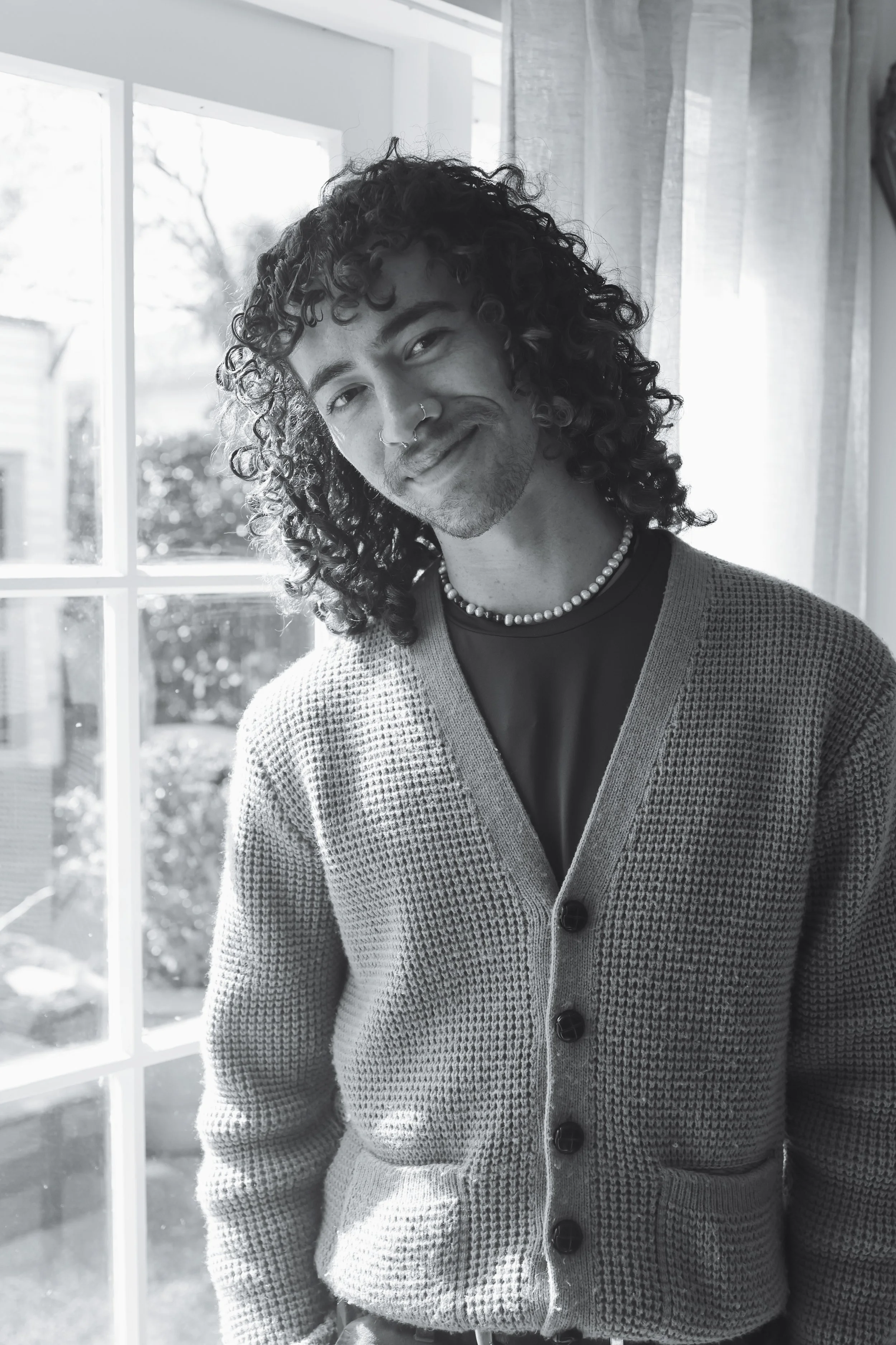 A person with curly hair wearing a pearl necklace and a cardigan standing by a window with sunlight shining through, smiling softly.
