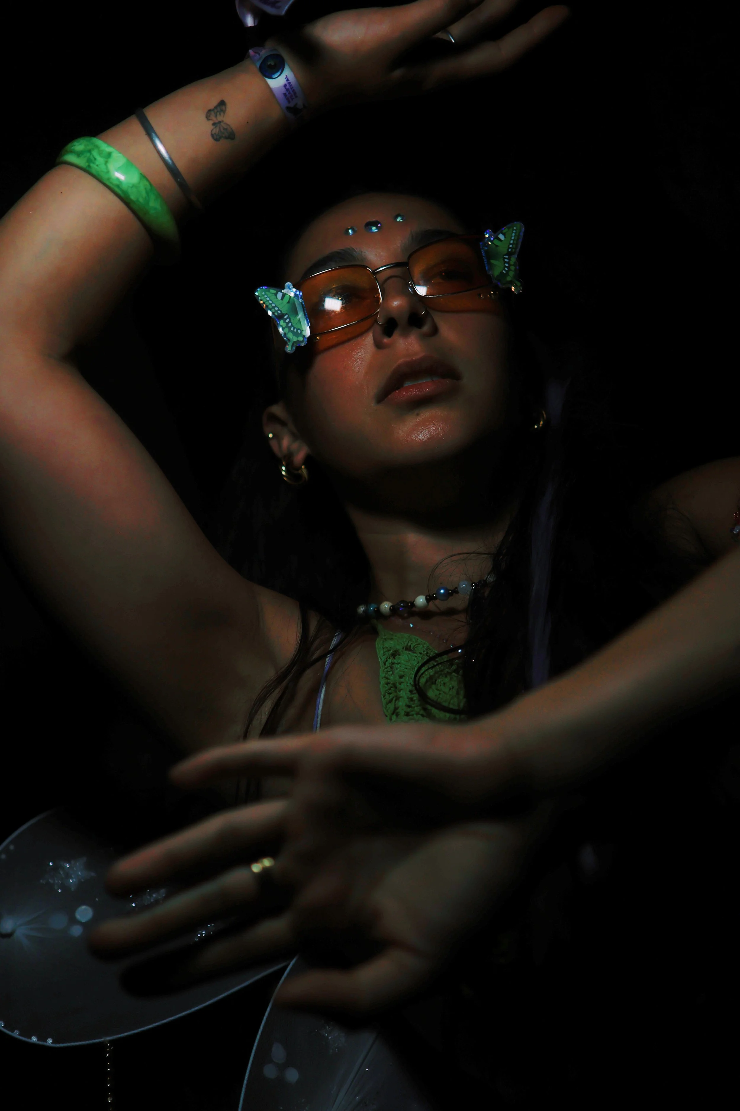 A woman lying down with her arm raised above her head, wearing sunglasses with butterfly-shaped decorations on the sides, a green top, and jewelry including earrings and a necklace. She has tattoos on her wrist, a bracelet, and a few decorative stones or dots on her forehead. The photo is taken with a mirror reflecting her face and part of her arm, with a dark background.