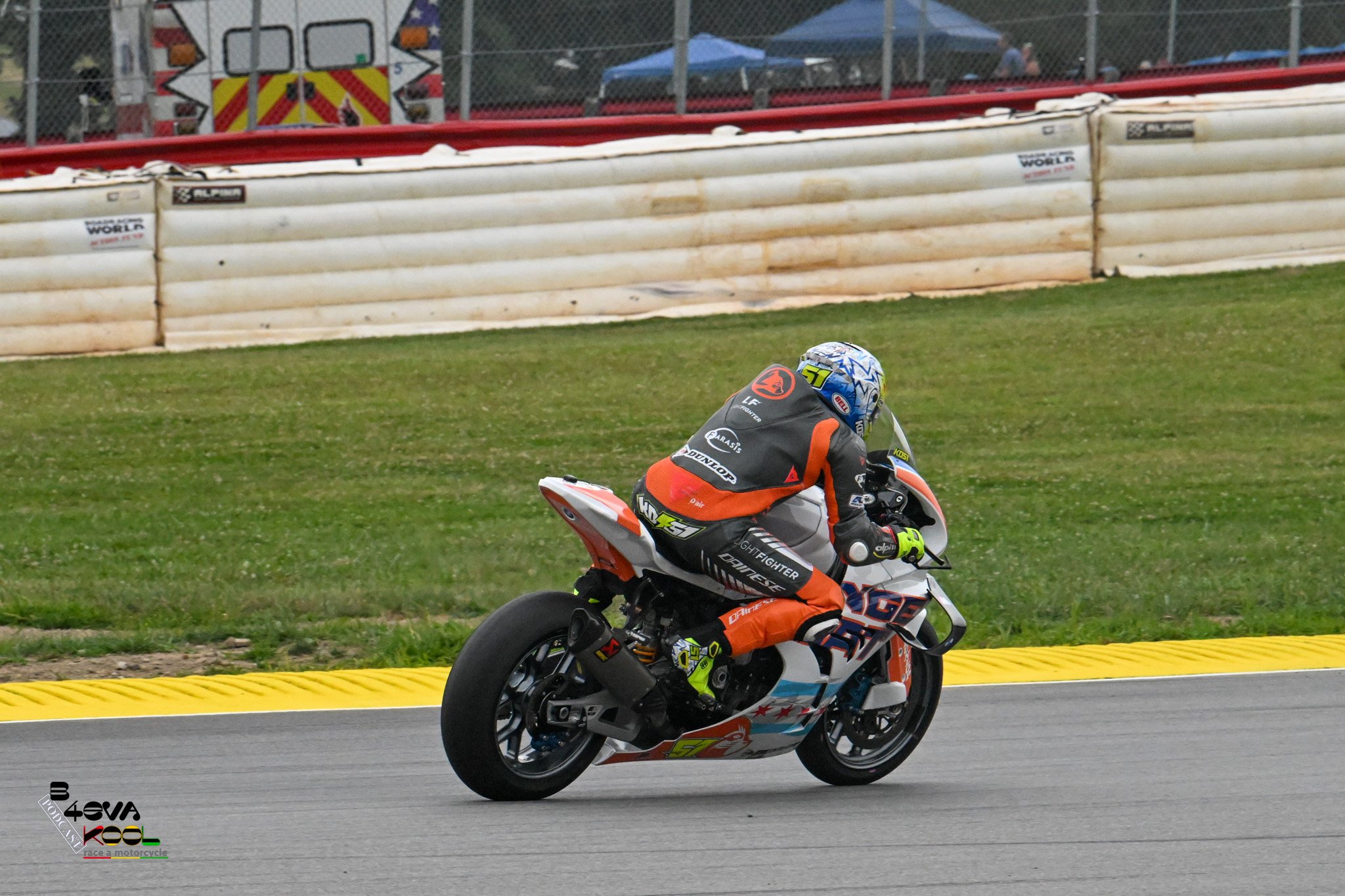 A motorcycle racer in orange and black gear riding on a racetrack with grass on the side. There are barriers and tents in the background at a racing event.