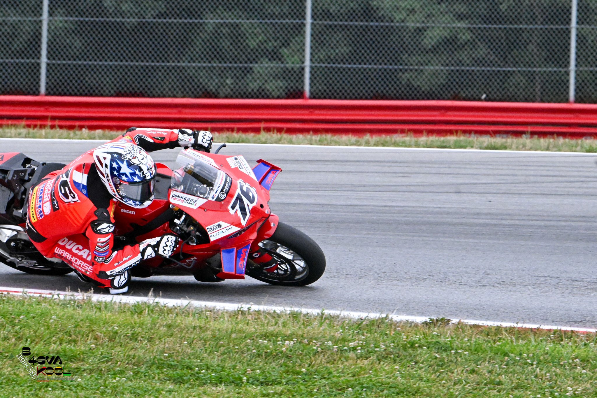 A motorcycle racer in a red racing suit and helmet leaning into a turn on a race track, with grass and red barriers behind.
