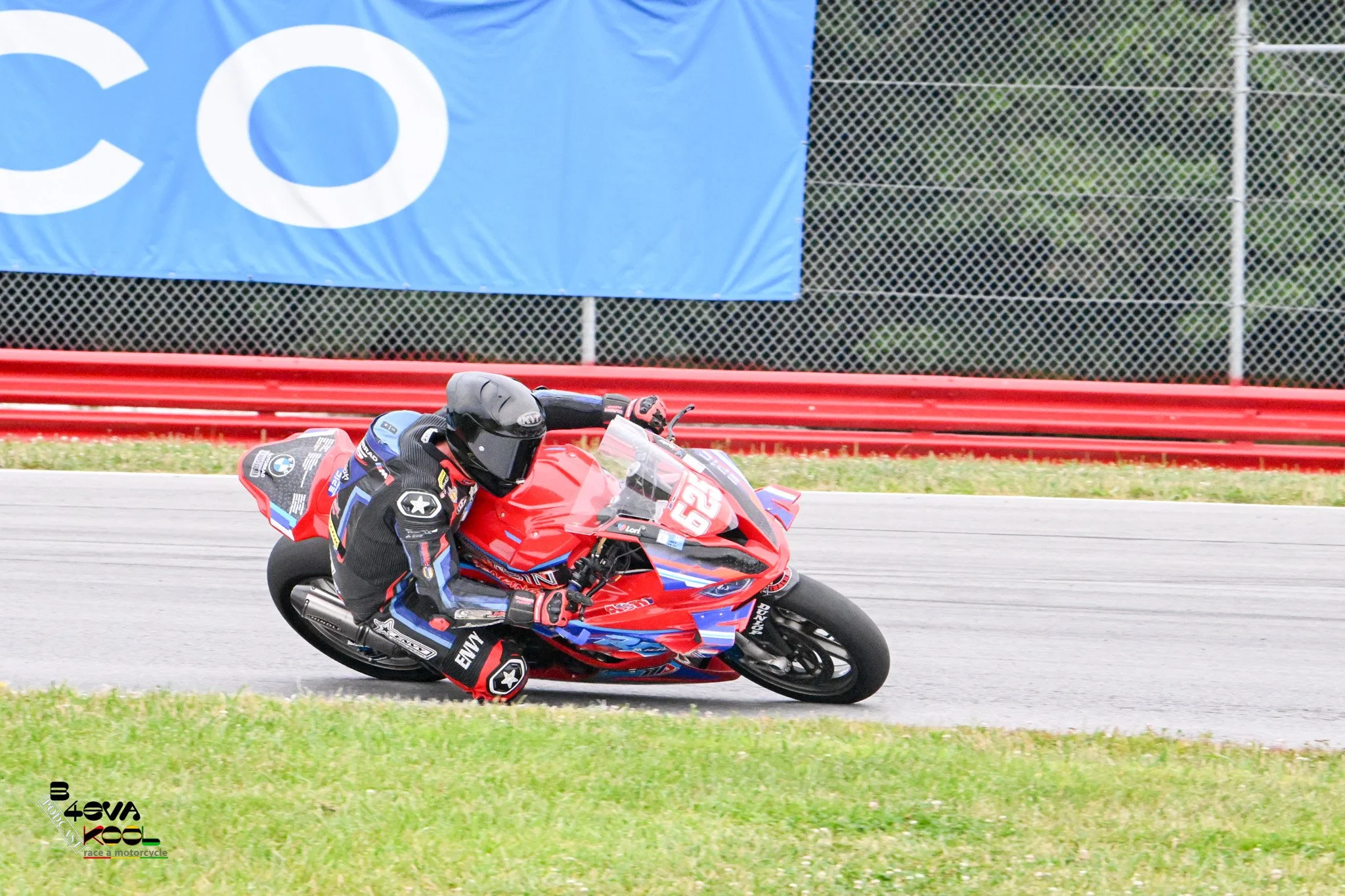 A motorcyclist wearing a black helmet and racing gear leaning into a turn on a red and blue motorcycle on a race track, with green grass, a chain-link fence, and a blue banner in the background.