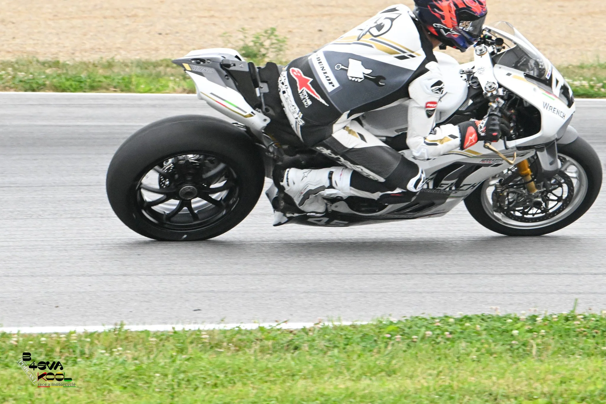 A motorcycle rider wearing a white racing suit and a red and black helmet riding a white sports motorcycle on a paved road with grass and dirt in the background.