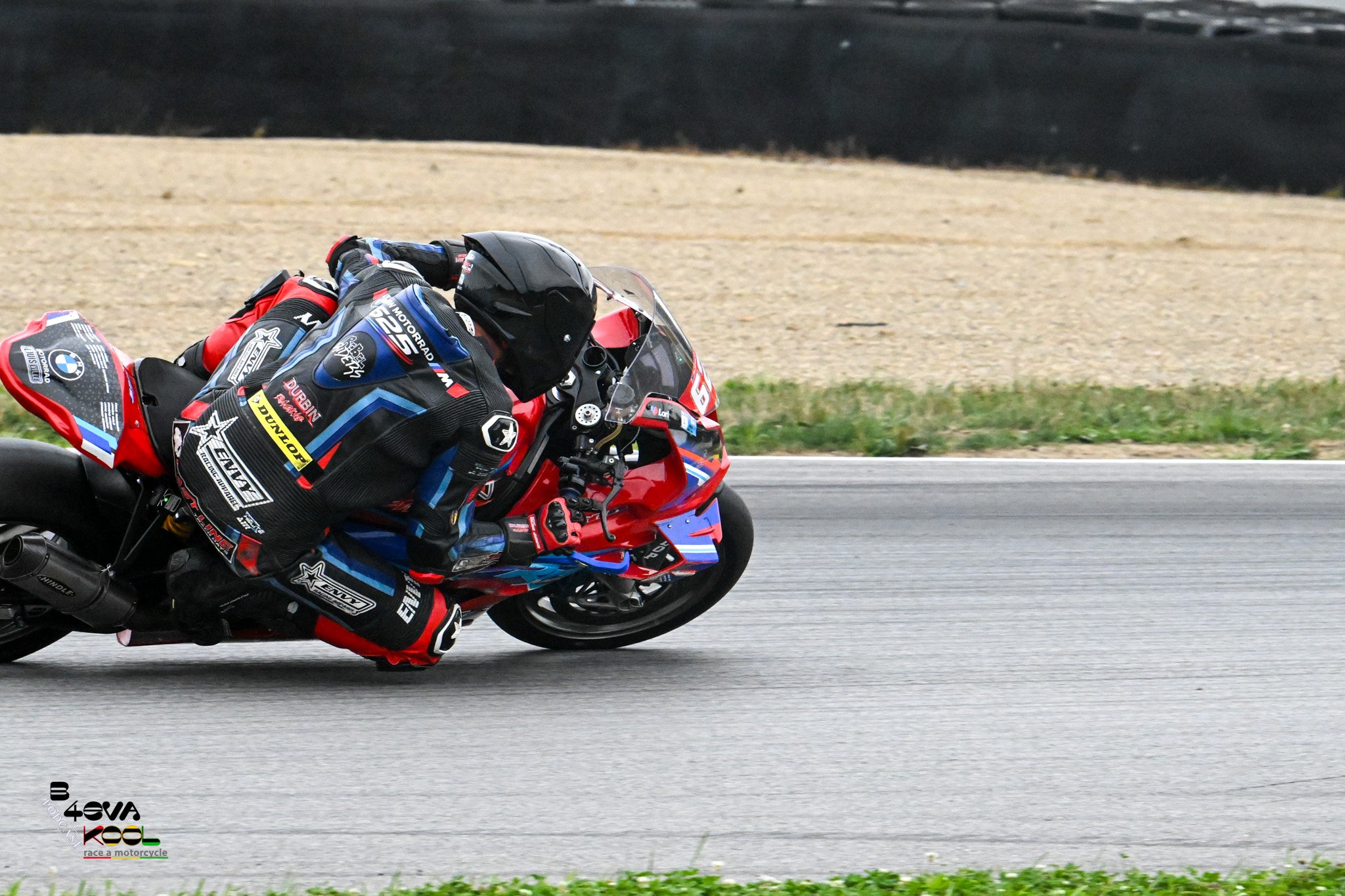 Motorcycle racer in black and red racing suit leaning into a turn during a race on a paved track with grassy area and gravel in the background.