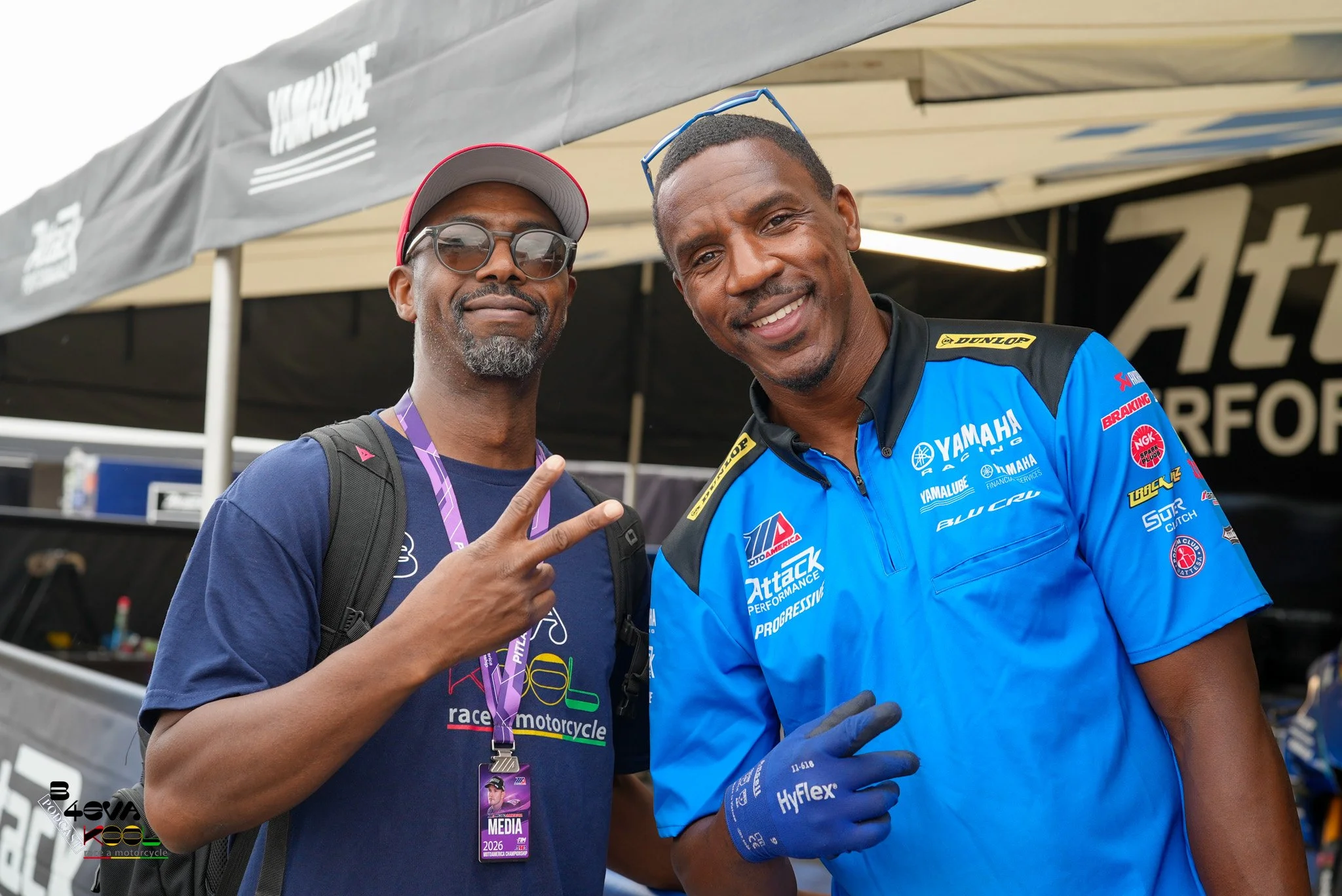 Two men posing together outdoors at a racing event. One is wearing a navy blue t-shirt, glasses, and an event pass around his neck, and the other is dressed in a blue Yamaha racing outfit with gloves. They are smiling and making peace signs.