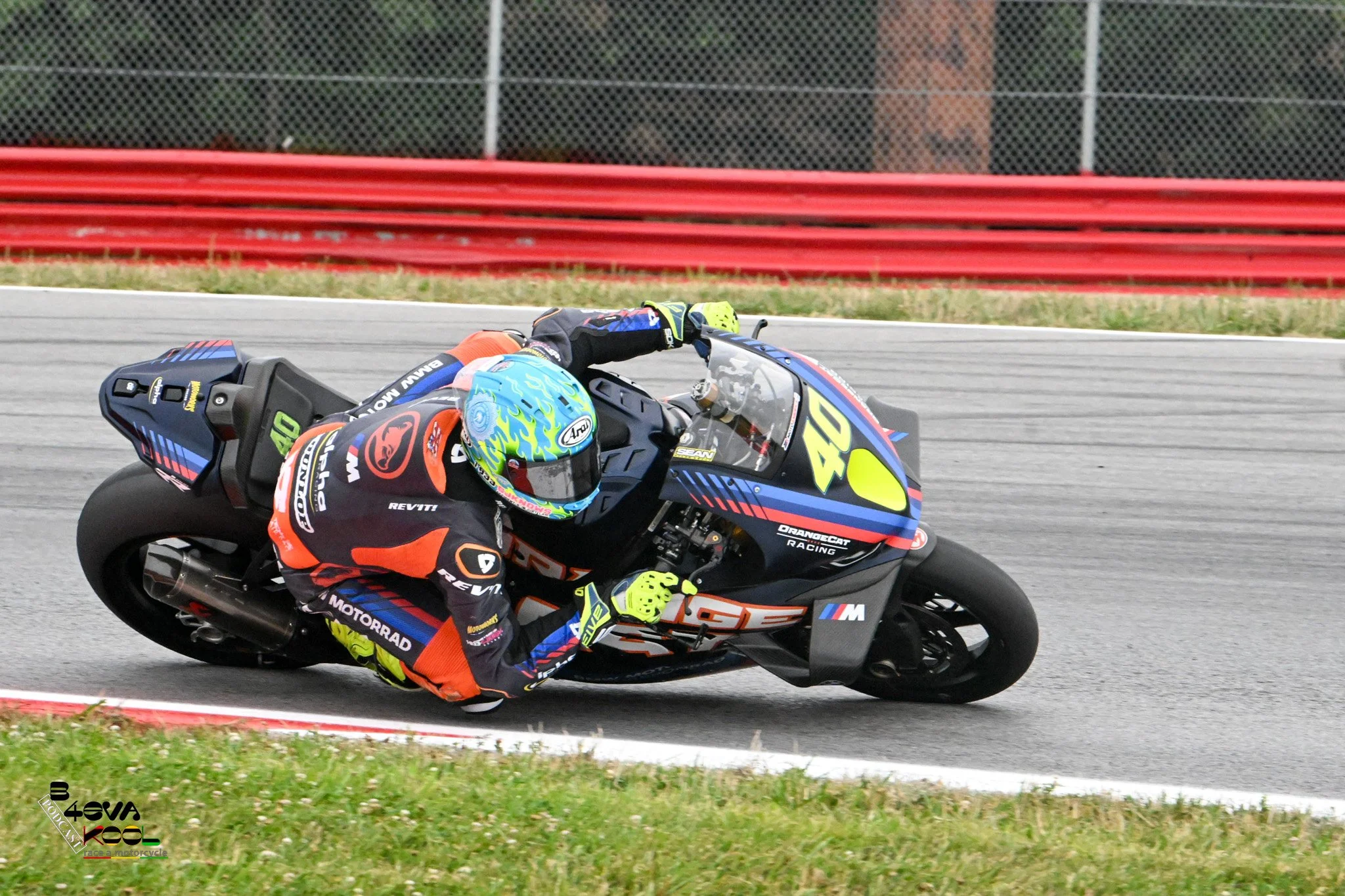 A motorcycle racer in colorful gear leaning into a turn on a racetrack with a red and green barrier in the background.