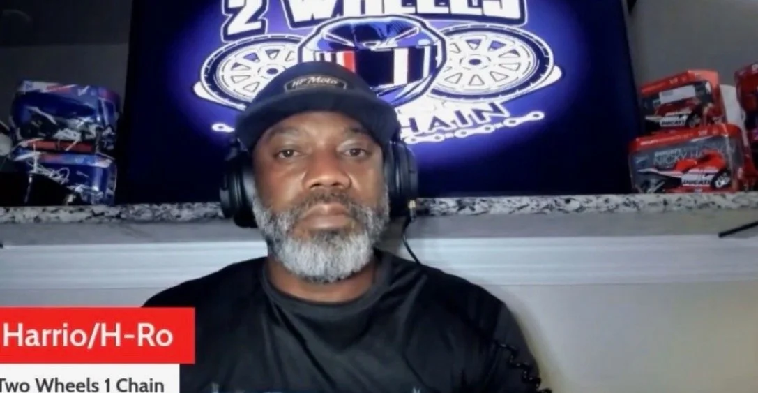 A man with a gray beard wearing a black hat and headphones, sitting in front of a television screen with a 'Two Wheels' motorcycle chain logo and several toy cars in packaging.