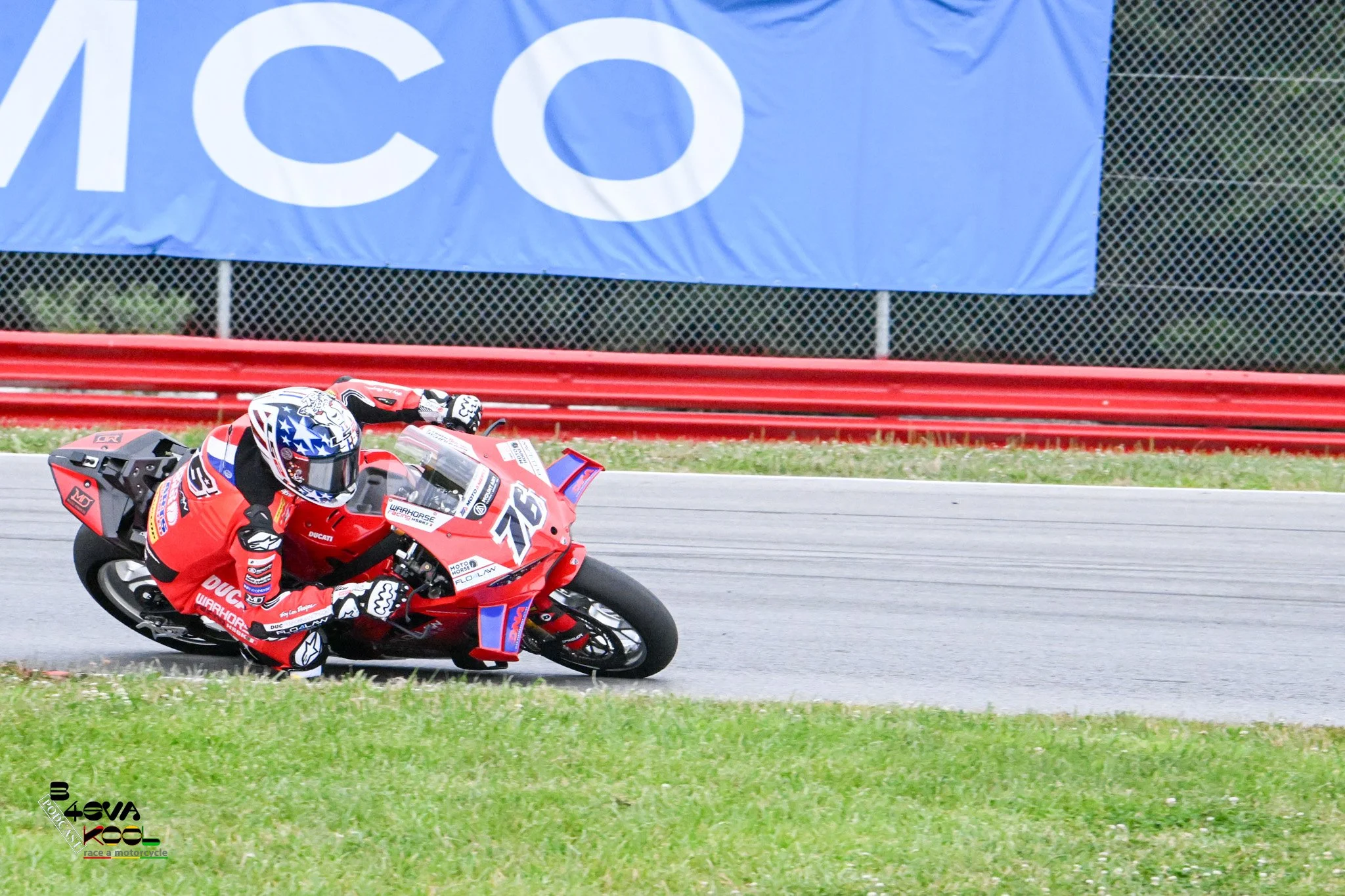 A motorcycle racer in red racing gear leaning into a turn on a race track, with a large blue banner and a red barrier in the background.