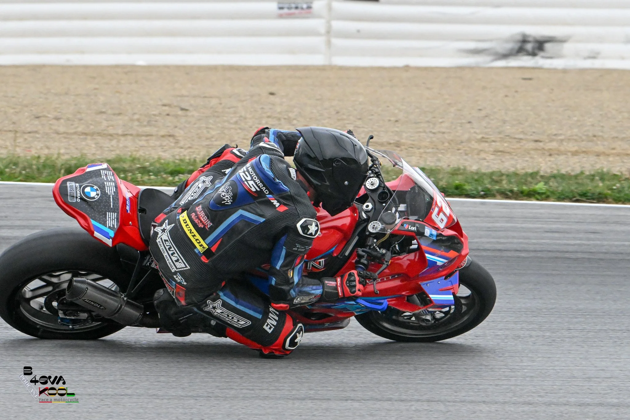 Motorcycle racer leaning into a turn on a racetrack, wearing red, black, and blue racing gear with sponsors' logos, and a black helmet.