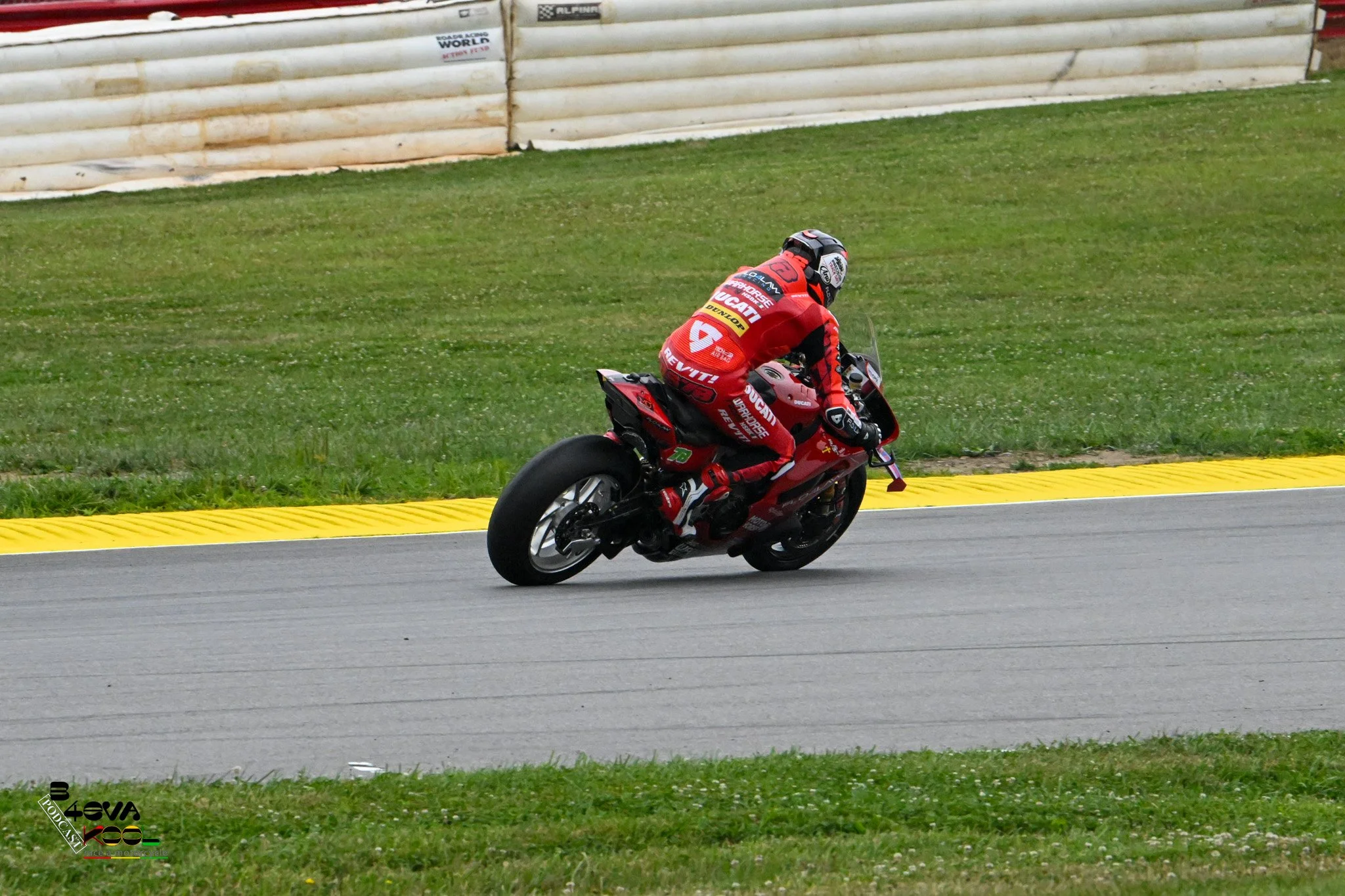 A motorcyclist in red racing gear and helmet leaning into a turn on a race track, with a grassy infield and a white barrier in the background.