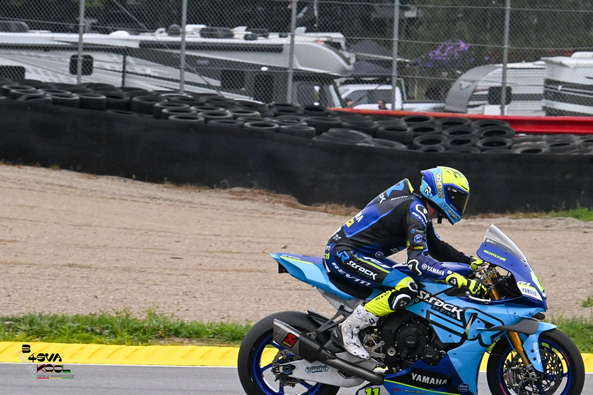 A professional motorcycle racer riding a blue Yamaha motorcycle on a racetrack, with tire stacks and fencing in the background.