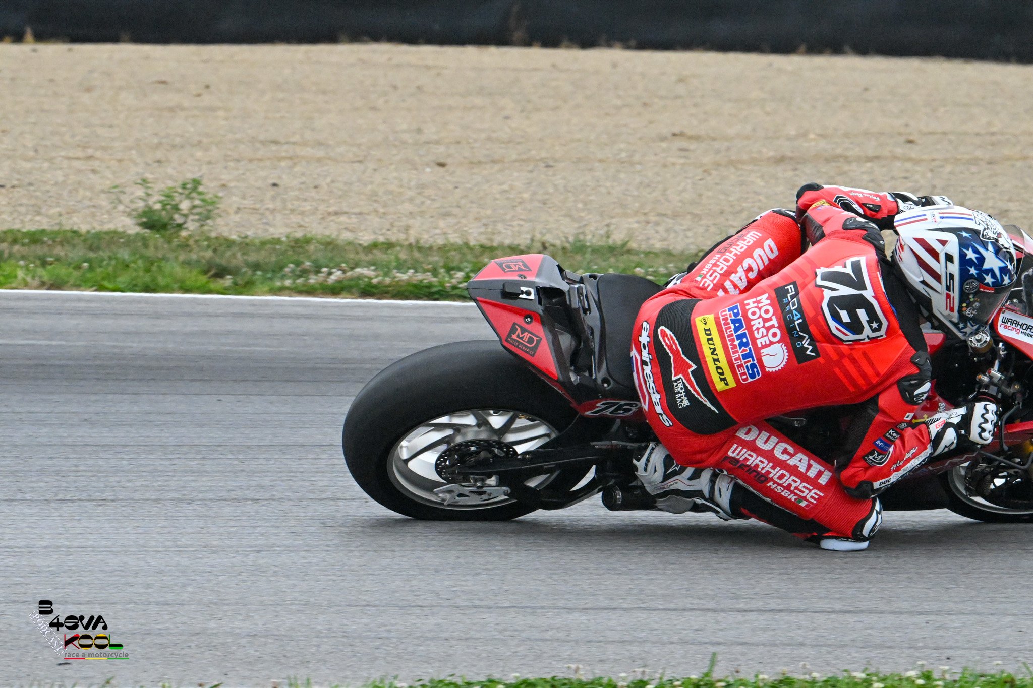 Motorcycle racer in red racing suit and helmet leaning into a turn on a racetrack, with grass and gravel visible in the background.