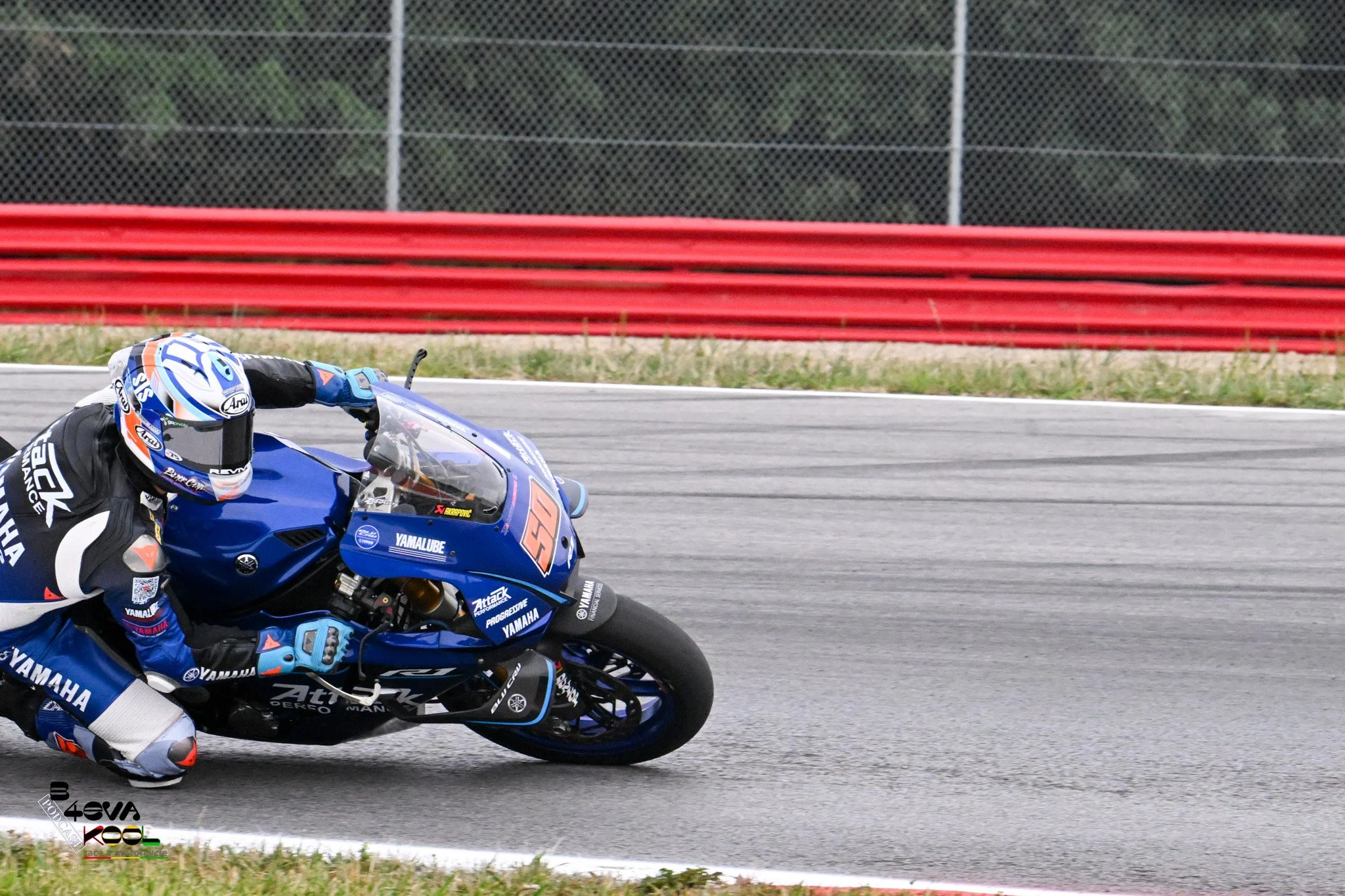 Motorcycle racer in blue racing suit leaning into a turn on a race track with red barriers and a chain-link fence in the background.