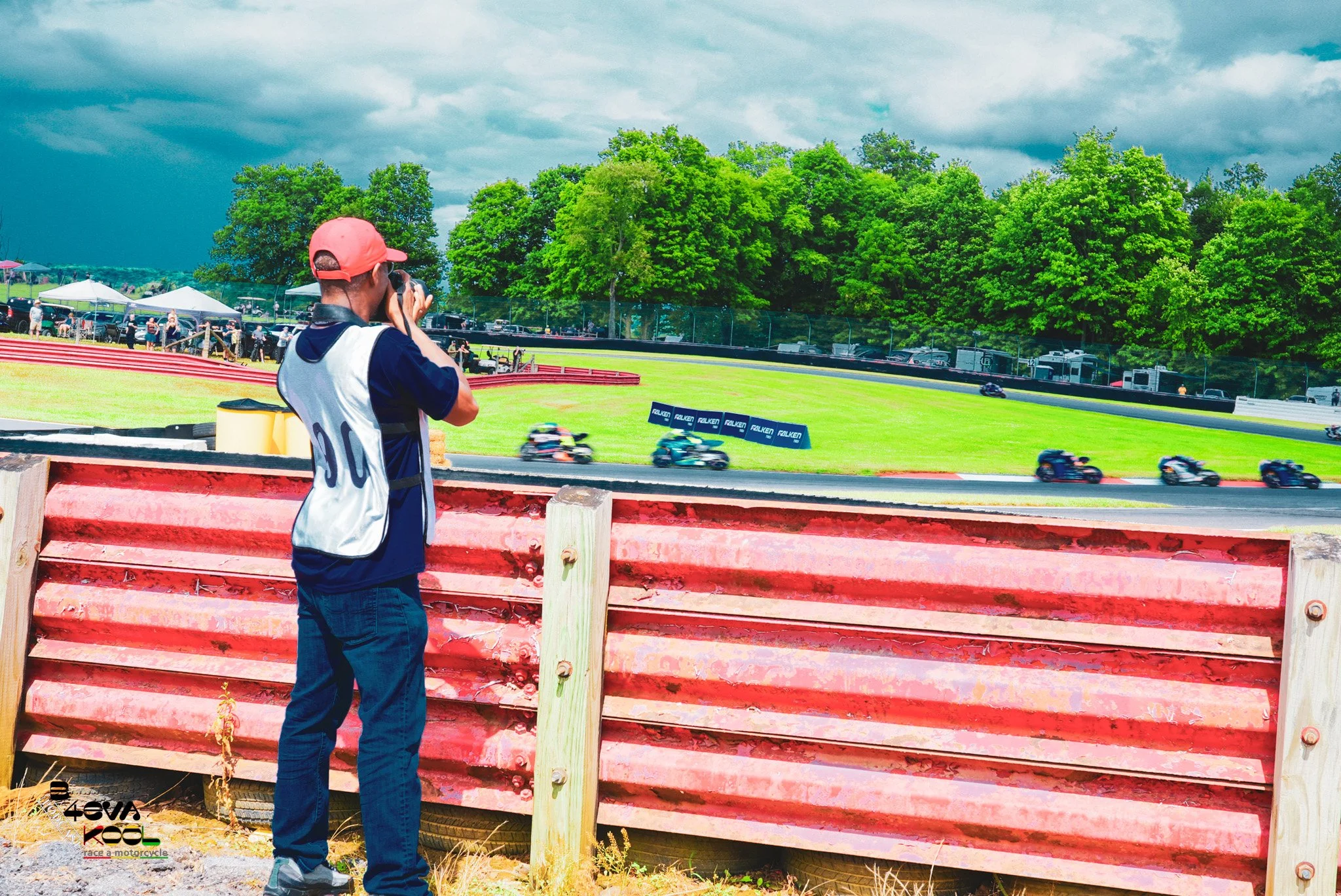 A person wearing a red cap and navy shirt taking a photo at a motorcycle race track with motorcycles speeding by the grass in the background, along with trees and a cloudy sky.